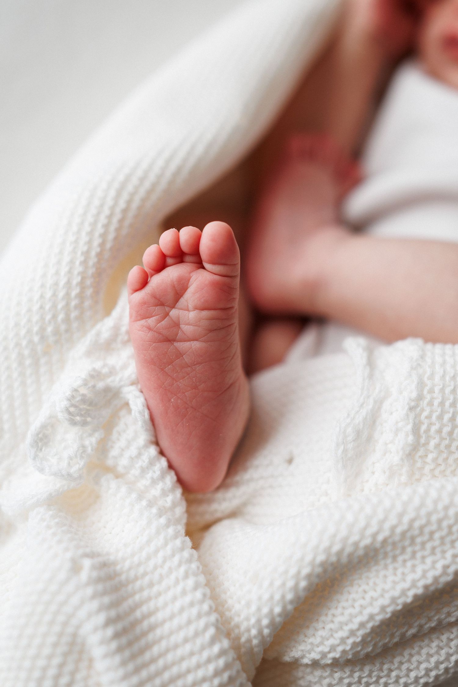 Close-up of a newborn baby's tiny bare foot resting on a soft cream knit blanket with the infant's hand softly blurred behind.