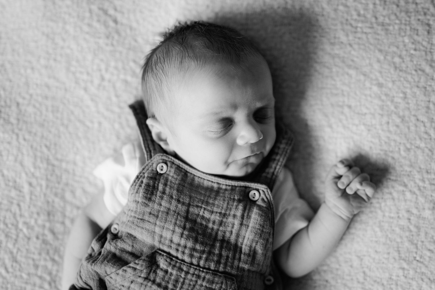 Black and white portrait of a newborn baby in muslin romper lying on a soft blanket.