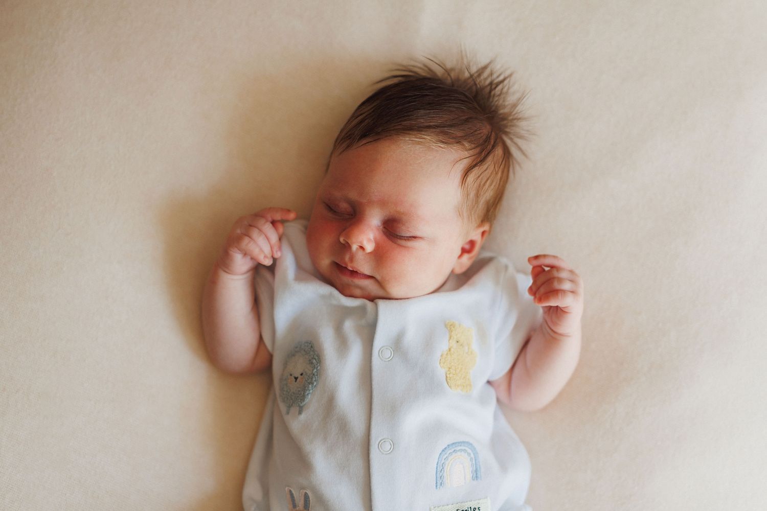Peaceful newborn baby sleeping on a cream blanket wearing a white sleepsuit with pastel rainbow and cloud appliques.