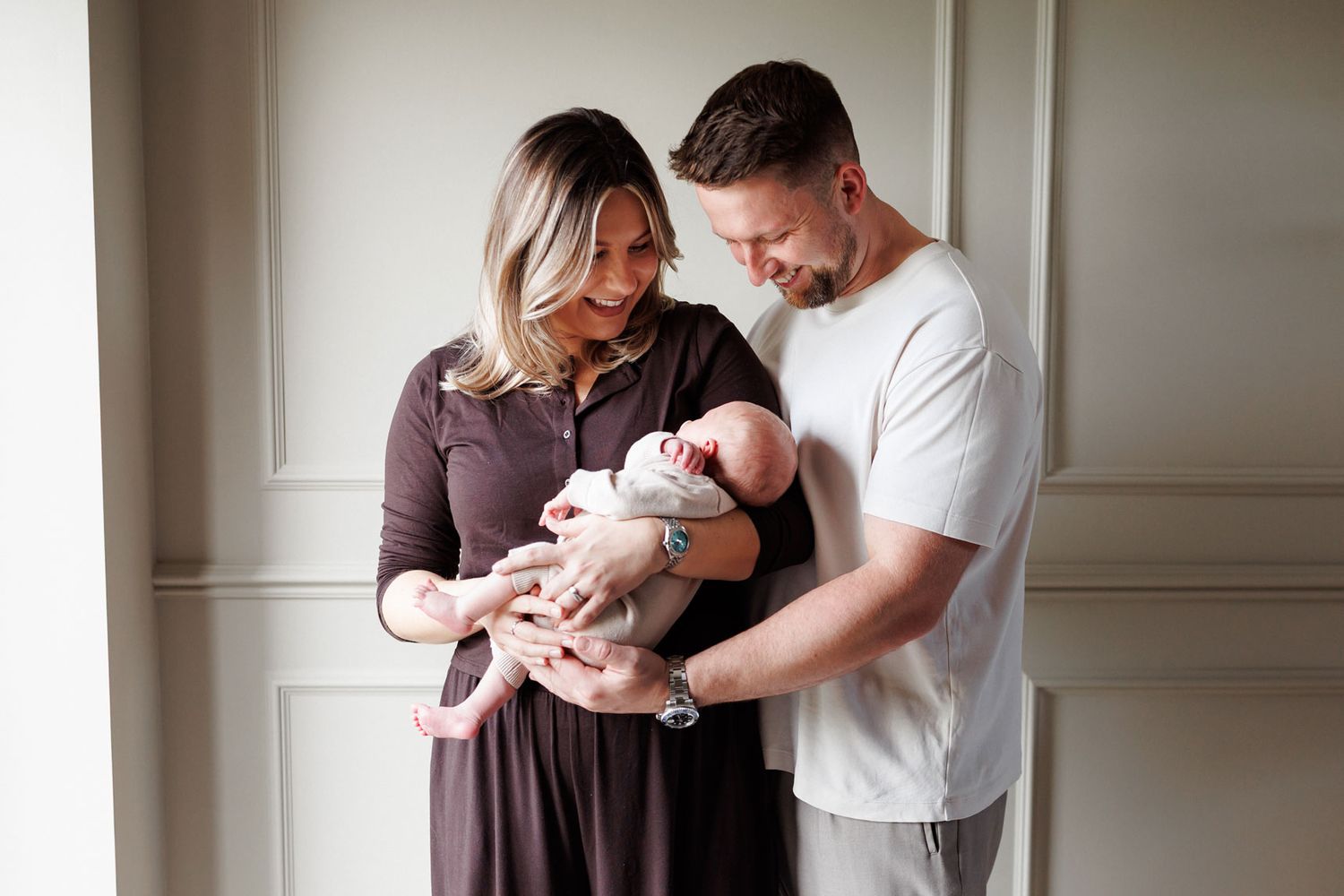 Smiling mother and father in casual clothes gently cradle their sleeping newborn baby during a bright indoor family portrait session.
