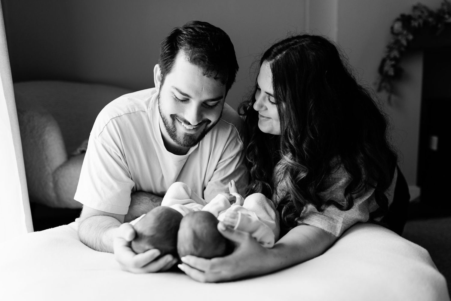 Black and white newborn photo of smiling parents gazing lovingly at their swaddled twin babies resting on a bed.