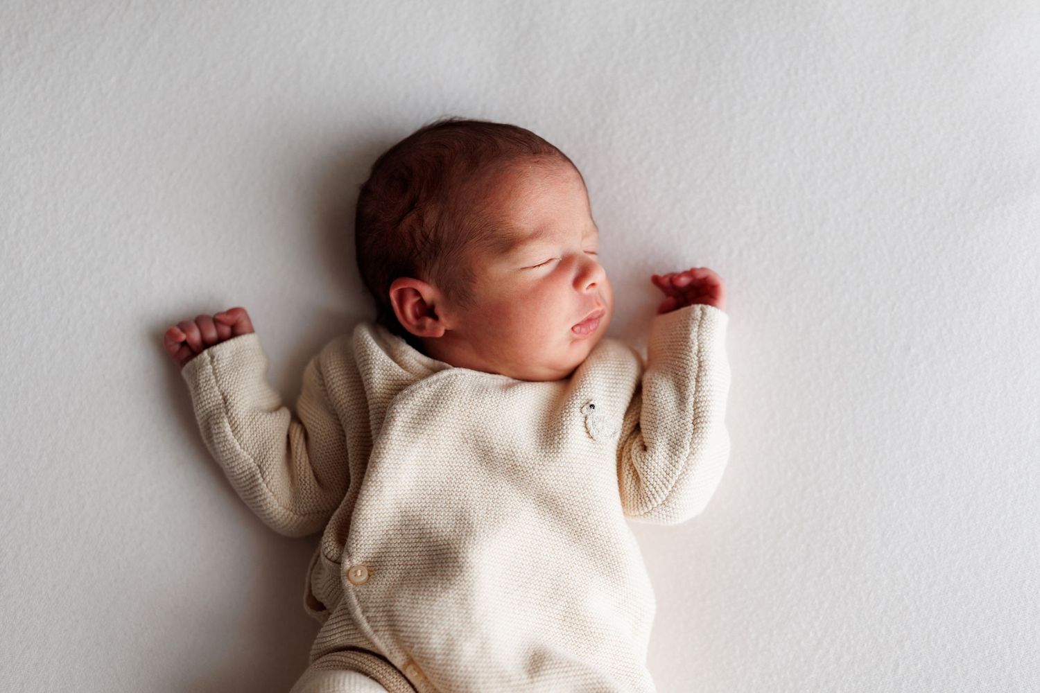 Sleeping newborn baby in a cream knit outfit lying on its back with arms raised on a soft white blanket.