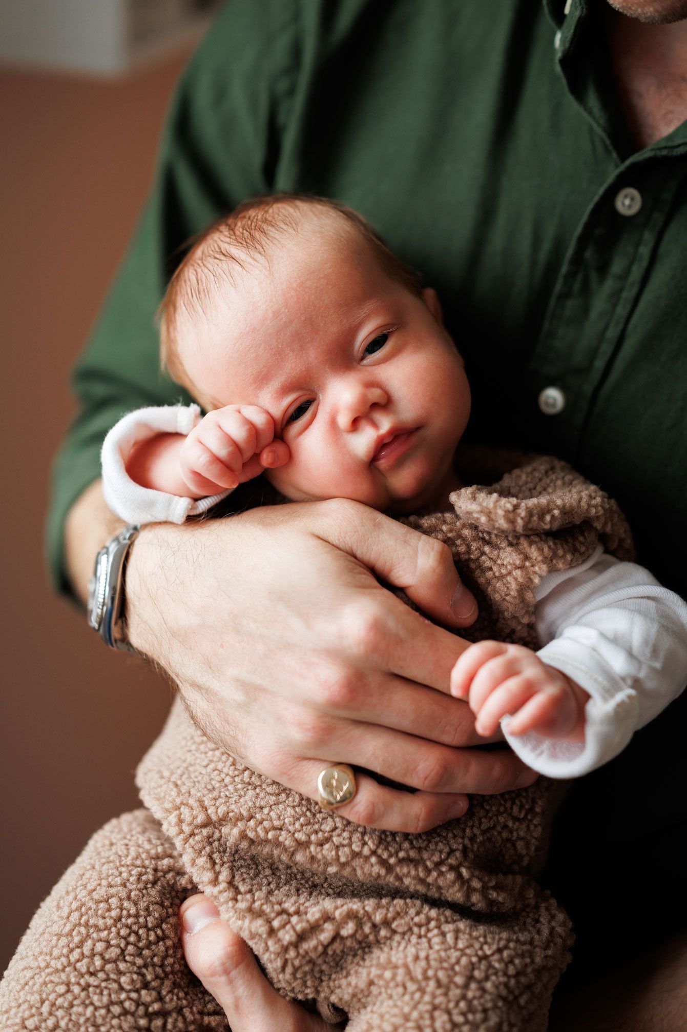 Newborn baby in a fuzzy brown sherpa outfit rests in the arms of a person in a green button-up shirt.