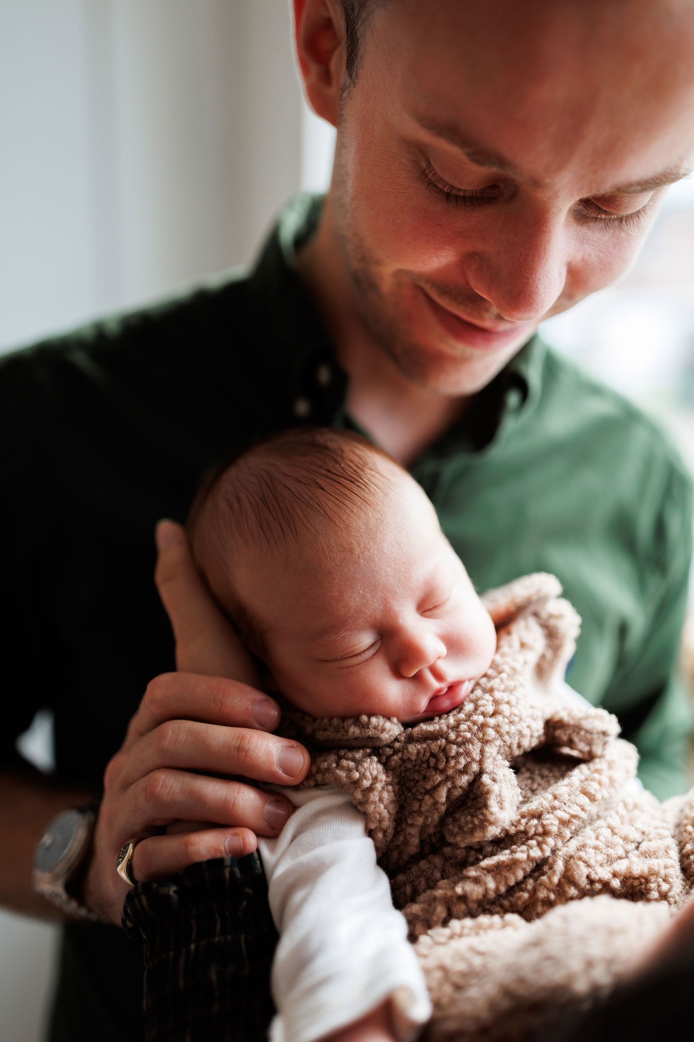 Smiling man in a green shirt gently cradles a sleeping newborn baby wearing a soft brown sherpa outfit.