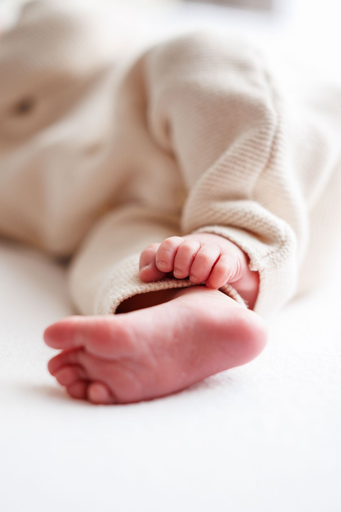 Close-up of a newborn baby's tiny bare foot and small curled hand resting on a soft white blanket, dressed in a cozy cream knit outfit.