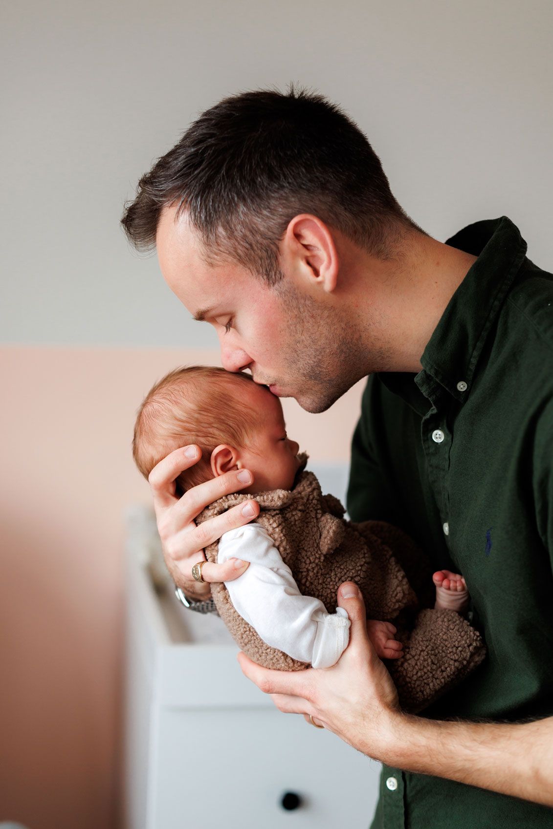 A father in a dark green shirt gently kisses the forehead of his newborn baby wrapped in a soft brown fleece outfit.