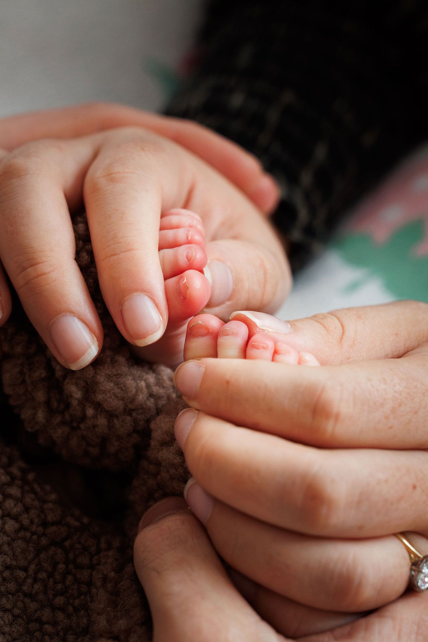 Two adult hands gently cradle a newborn baby's tiny feet against a soft brown fleece blanket.