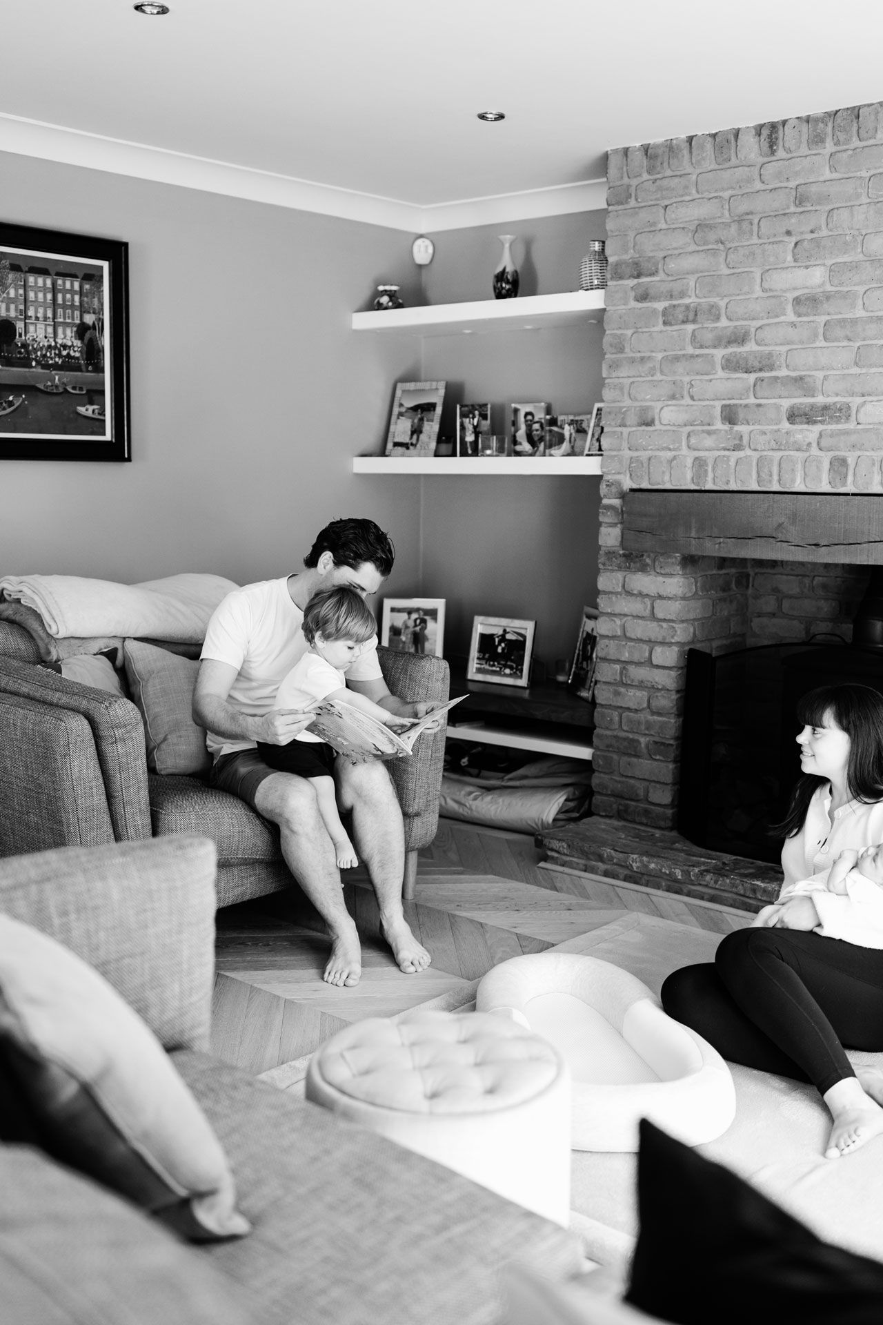 Black and white photo of a father reading to his toddler on a sofa while a woman sits on the floor by a brick fireplace.