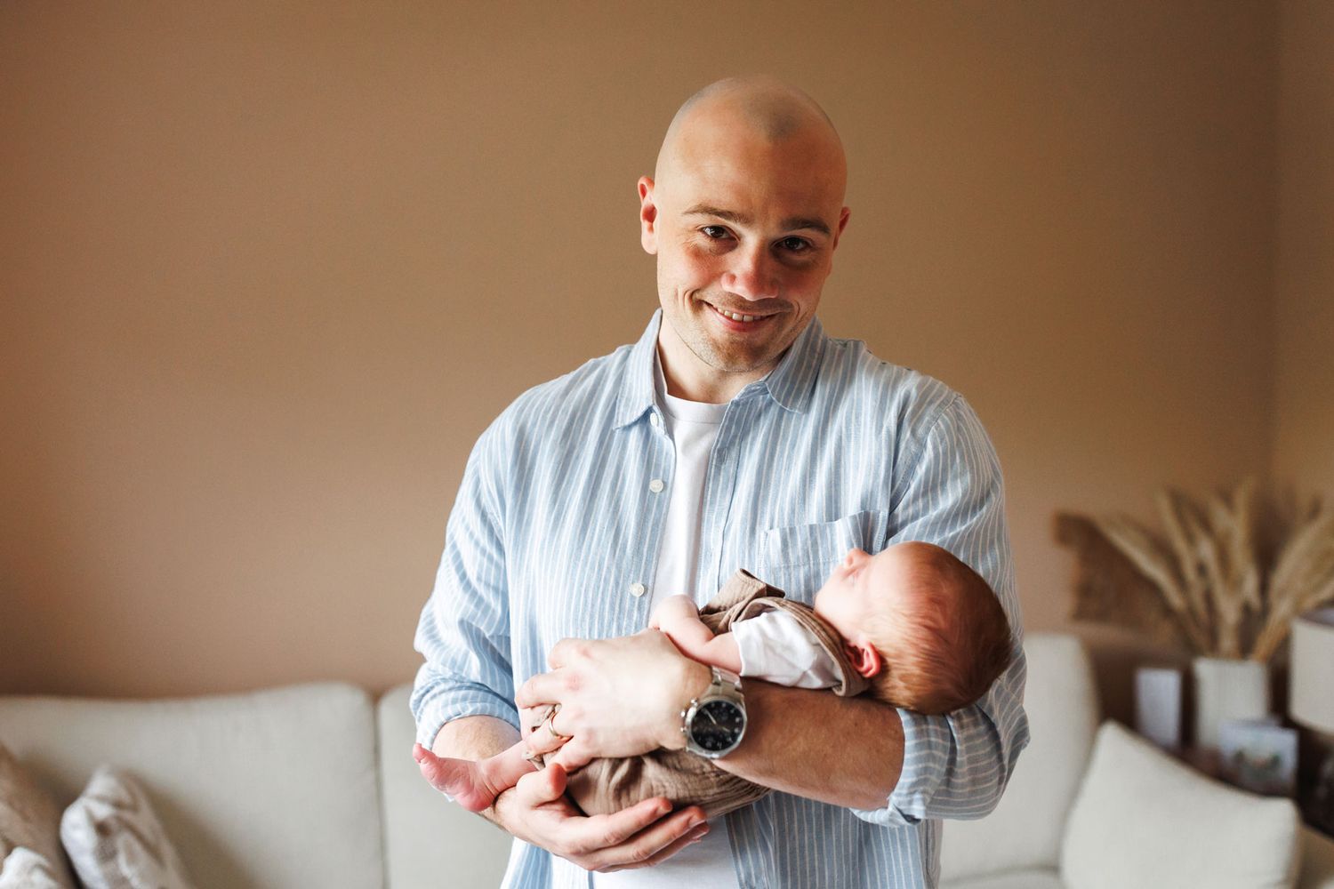 Smiling bald man holding a newborn baby in his arms, standing in a cozy living room wearing a blue striped shirt.
