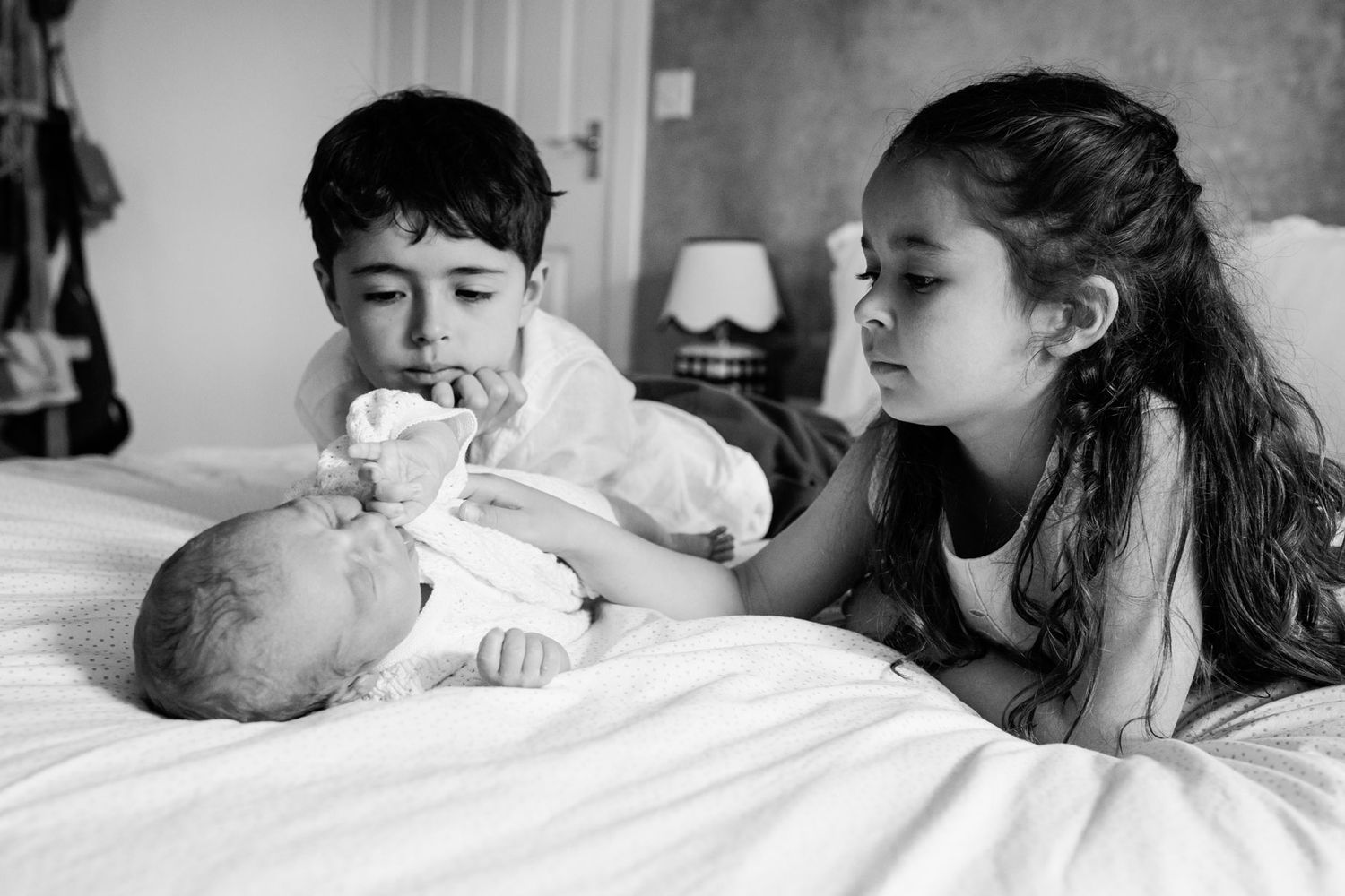 Two older siblings tenderly gaze at their newborn baby on a bed in black and white photo.