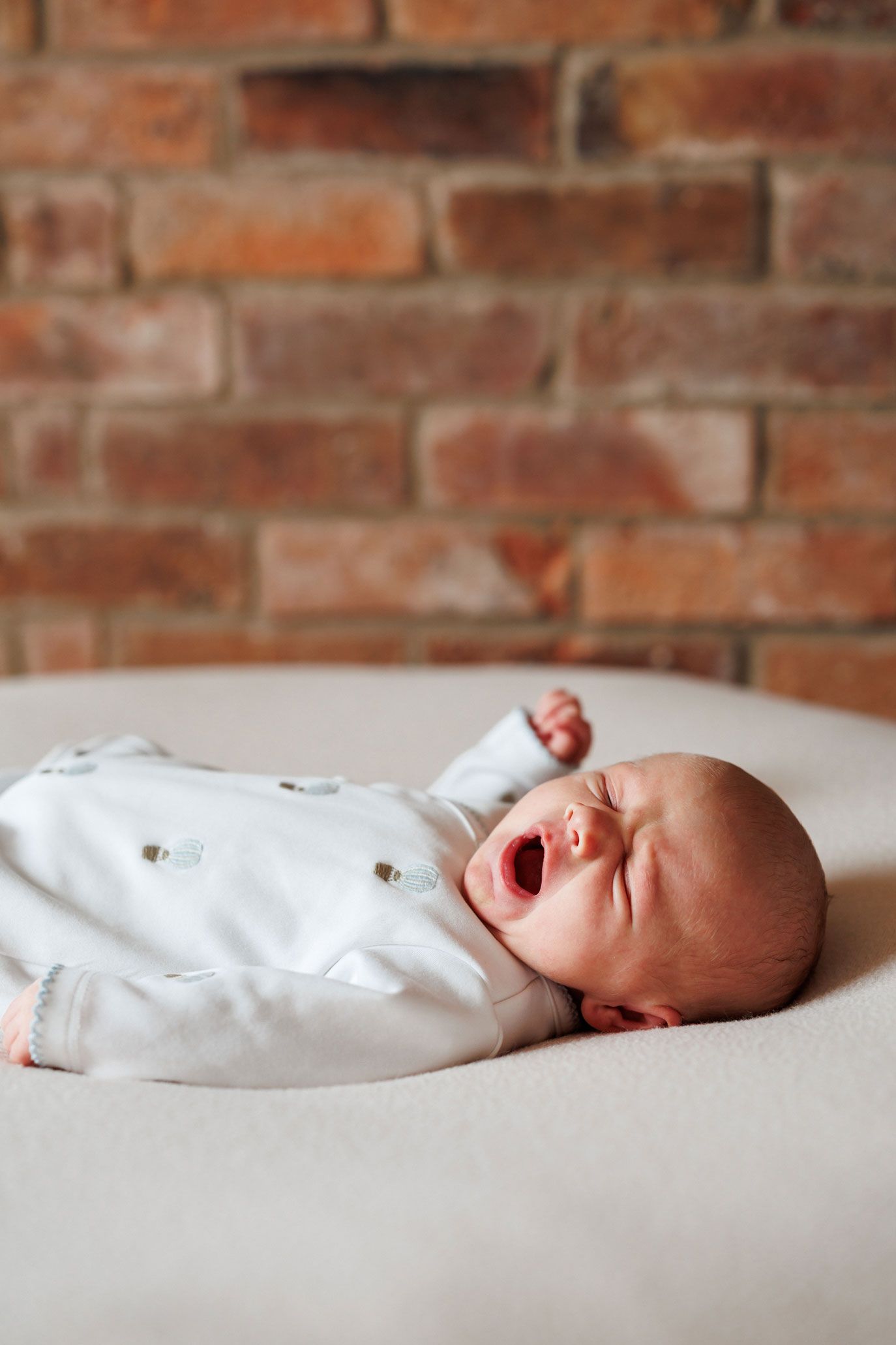 Newborn baby in a white long-sleeved outfit yawning while lying on a soft cream blanket in front of a rustic red brick wall.