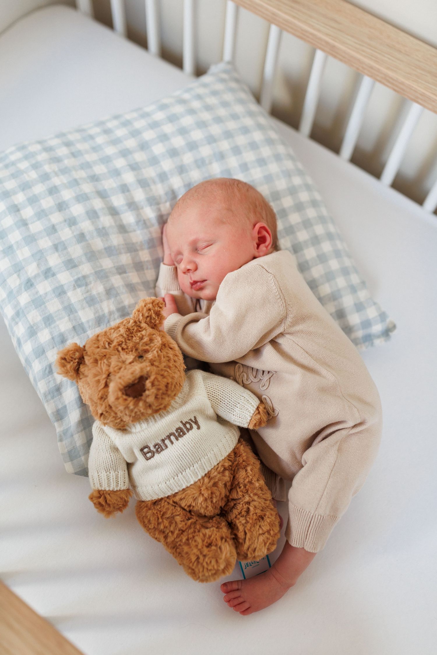 Sleeping newborn baby in a cream outfit cuddling a plush teddy bear in a white sweater reading Barnaby on a blue gingham pillow in a crib.