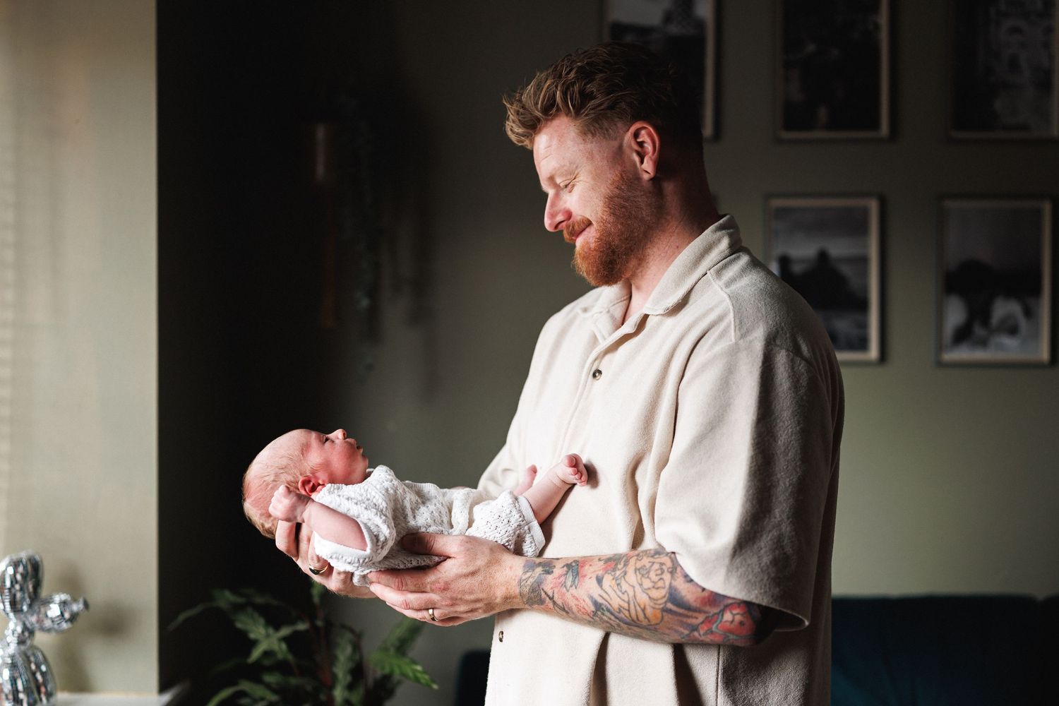 Tattooed red-haired father gently holding his sleeping newborn baby in a cozy home setting.