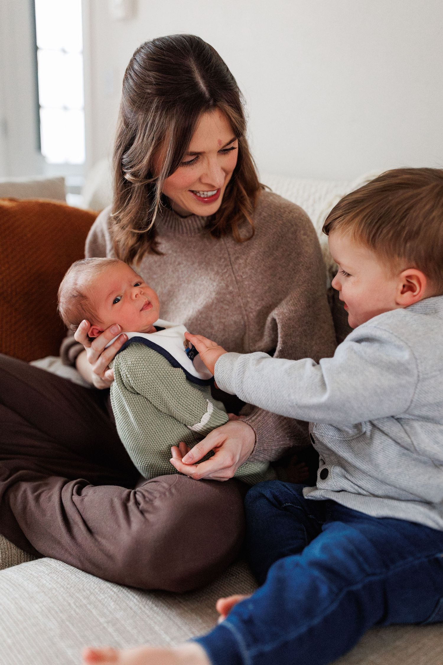 A mother on a couch carefully holds her newborn baby while her young toddler son reaches over to touch it.