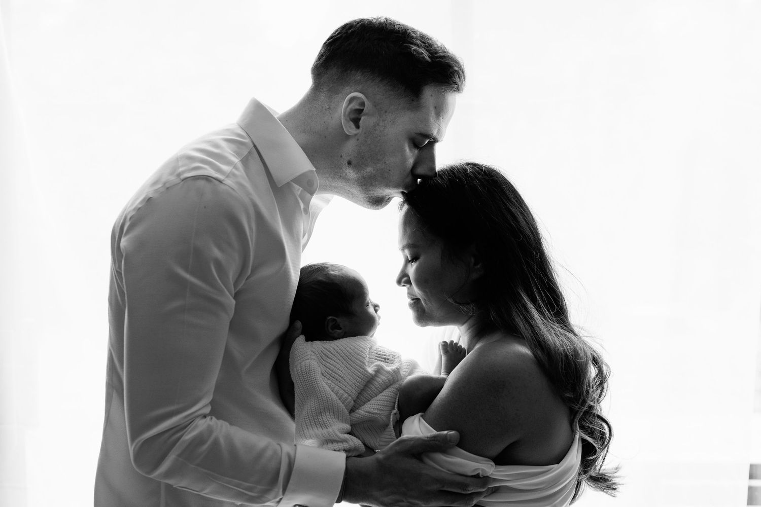 Black and white portrait of parents tenderly holding their newborn baby by a bright window.