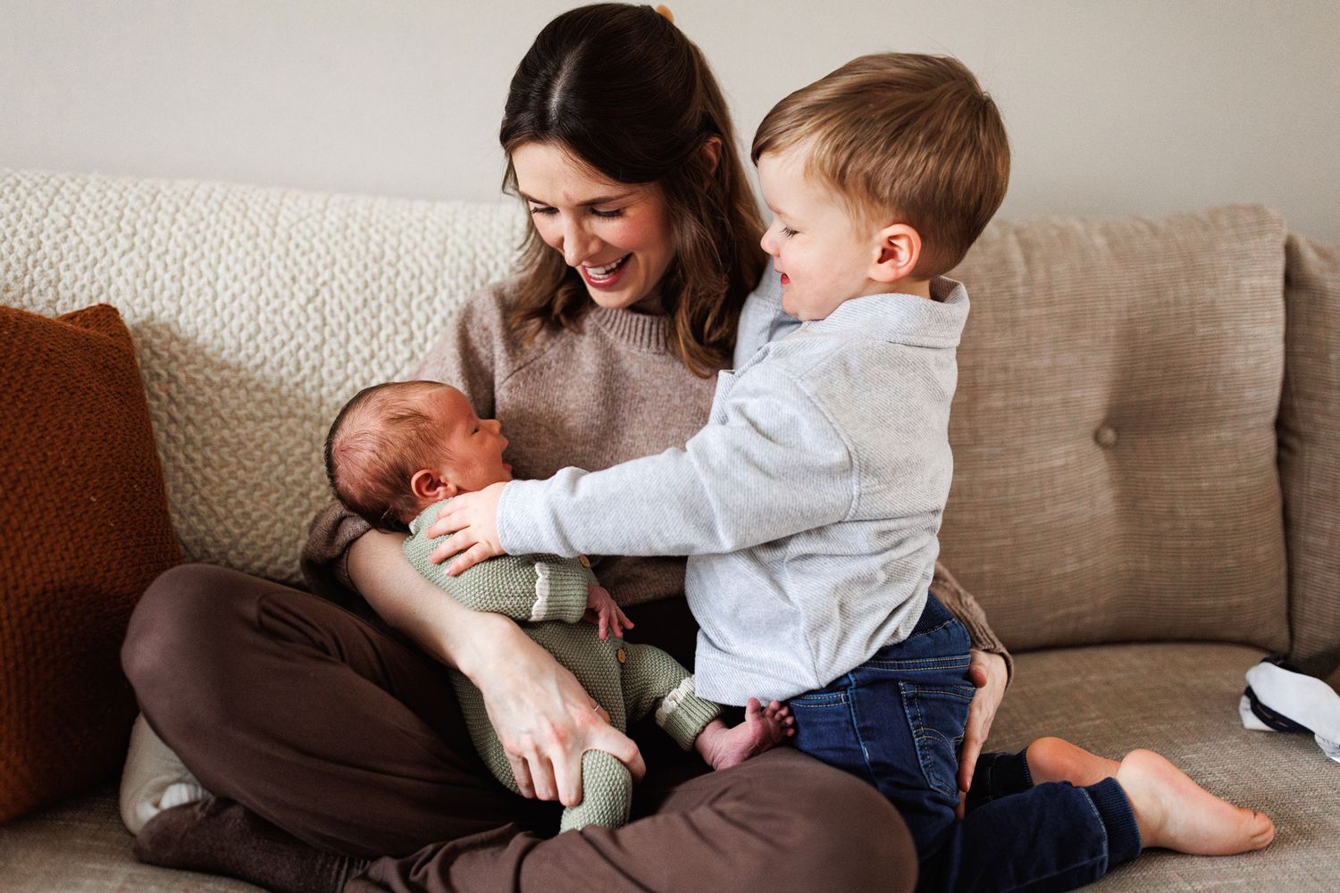 Smiling mother sits cross-legged on a sofa holding a newborn while a toddler reaches over to touch the baby.