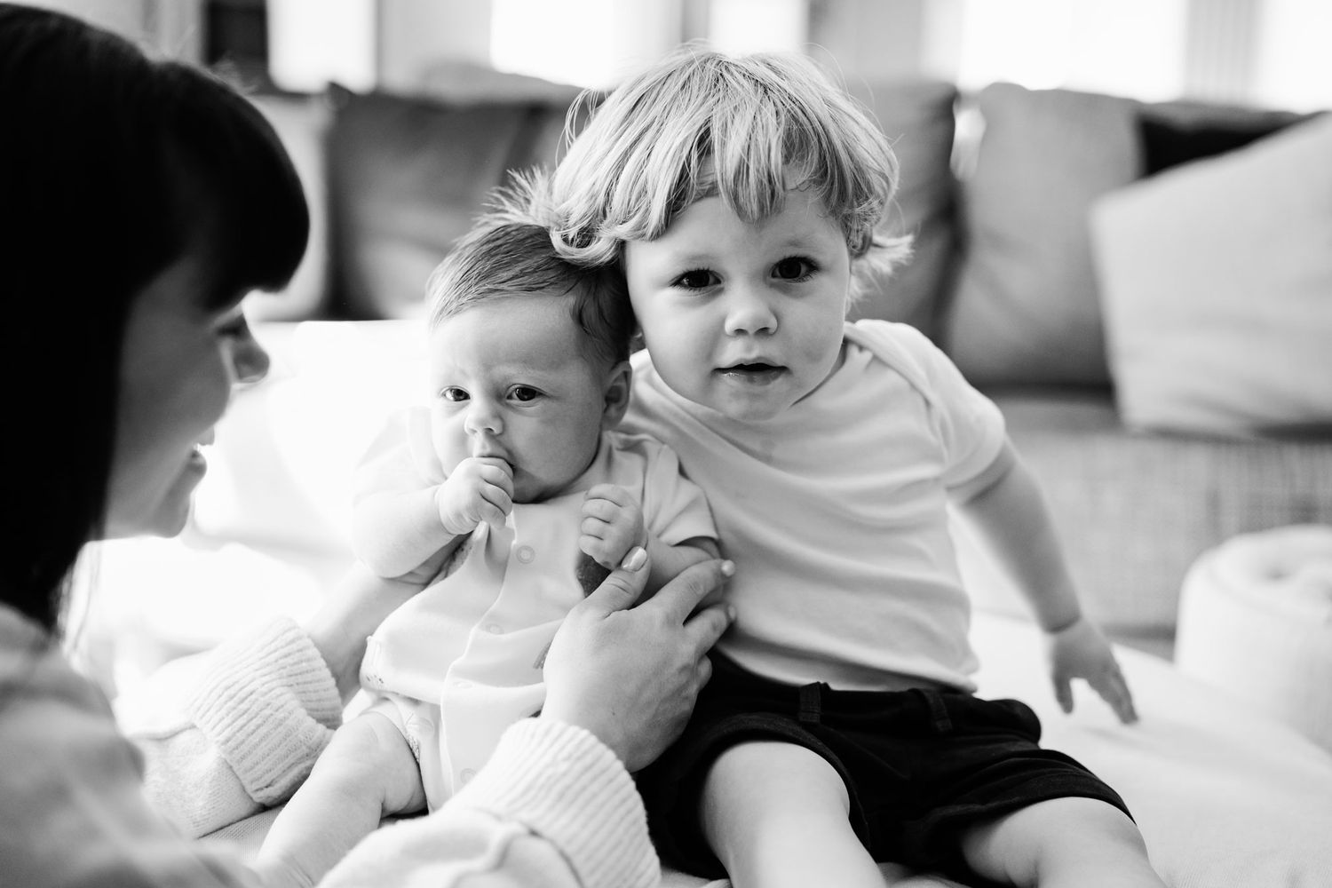 Black and white photo of mother holding two young children, toddler boy and baby, on a couch indoors.