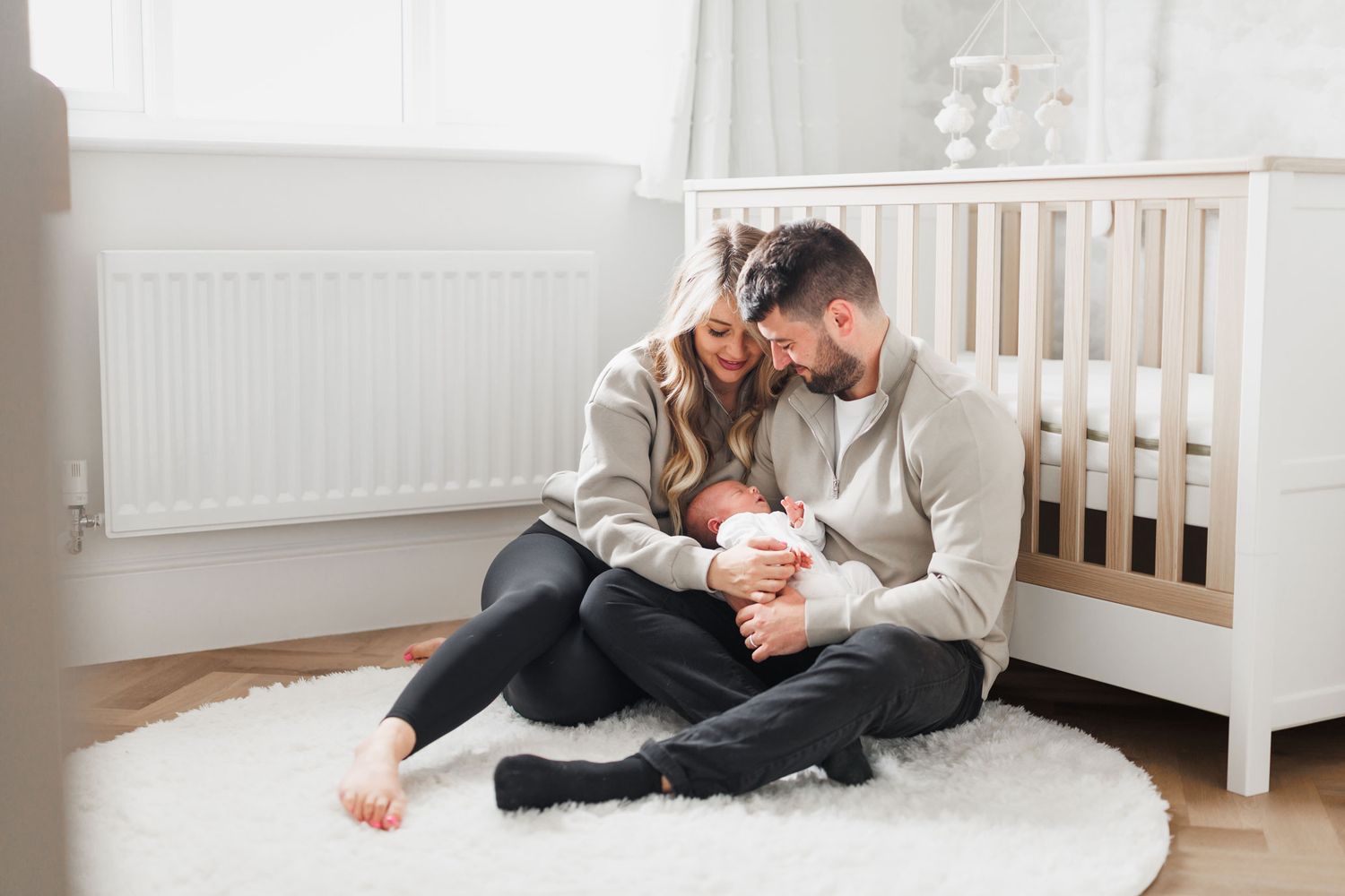 Happy couple in matching grey sweatshirts sitting on a round rug cradling their newborn baby by a white crib.