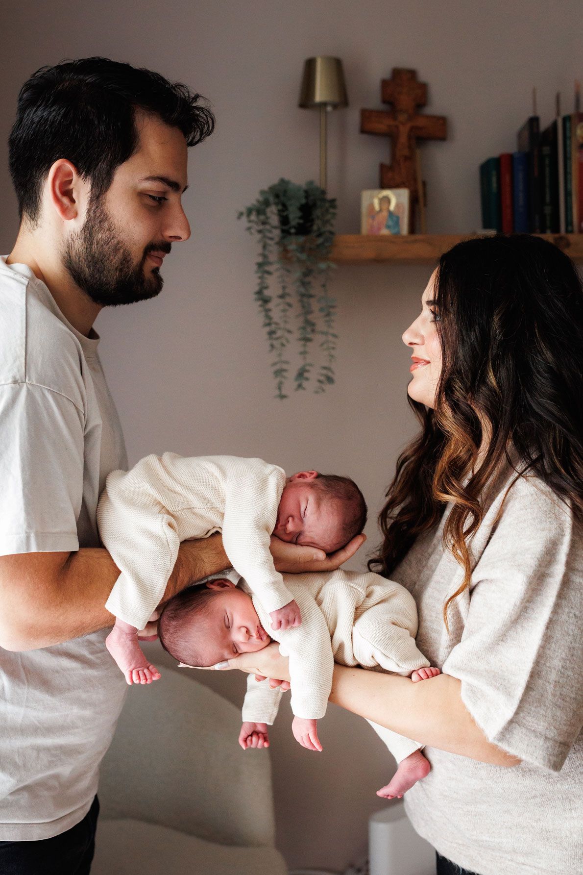 Smiling parents hold two sleeping newborn twins in cream outfits in a cozy room with a wooden cross and hanging plant.