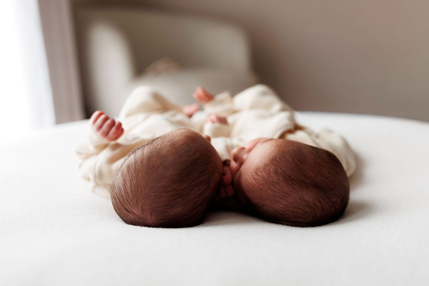Two newborn twin babies in cream onesies lie side by side on a soft white bed, their heads touching and tiny feet visible.