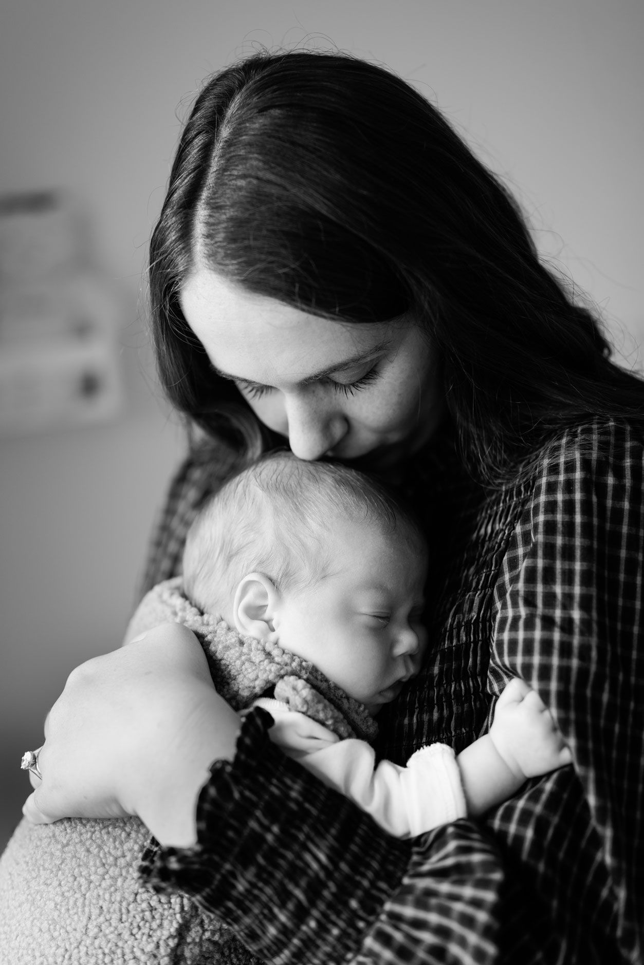 Black and white portrait of a mother with long dark hair tenderly kissing her sleeping newborn baby while holding the infant close.