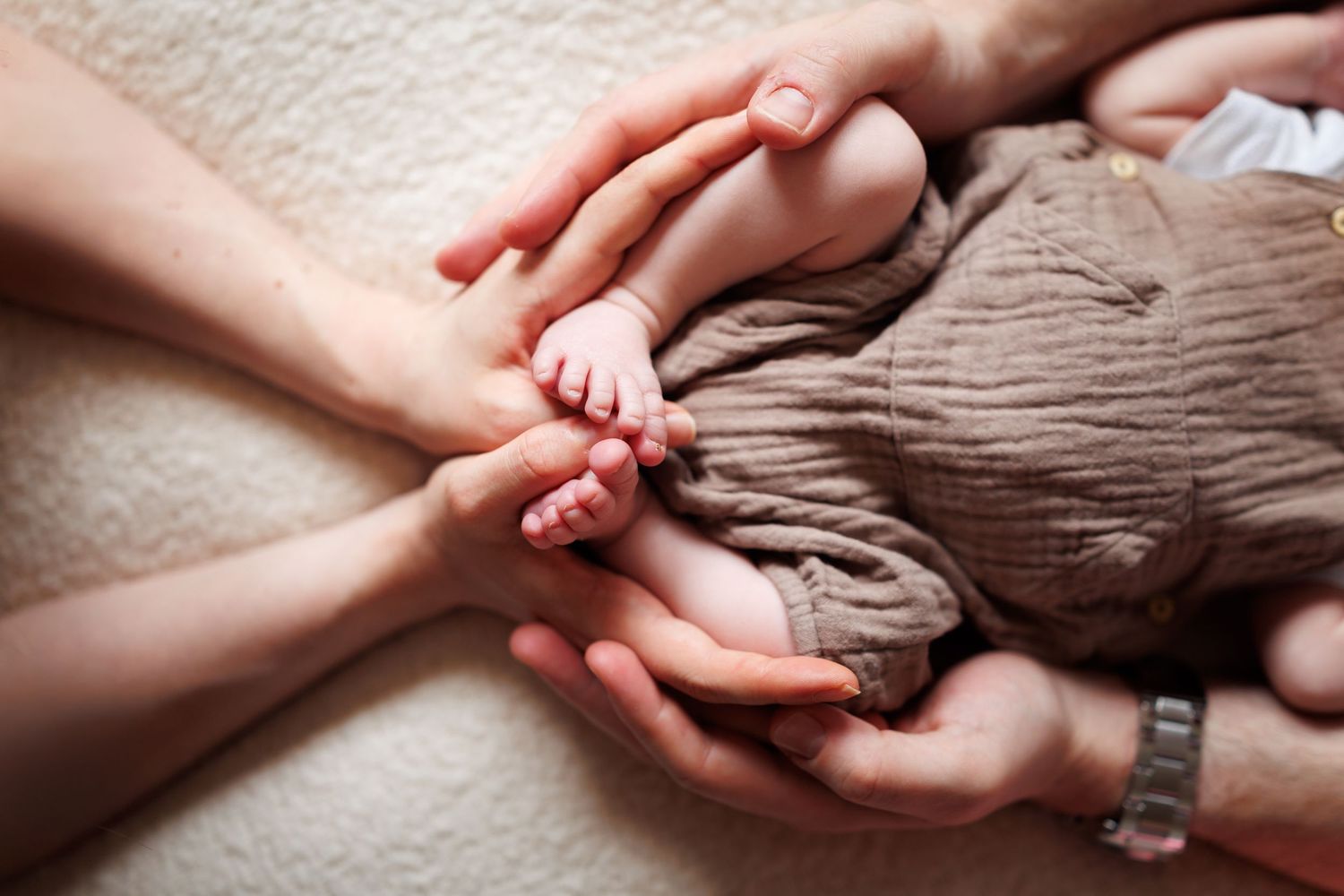 Parents gently holding their newborn baby's tiny feet and hands on a soft blanket.