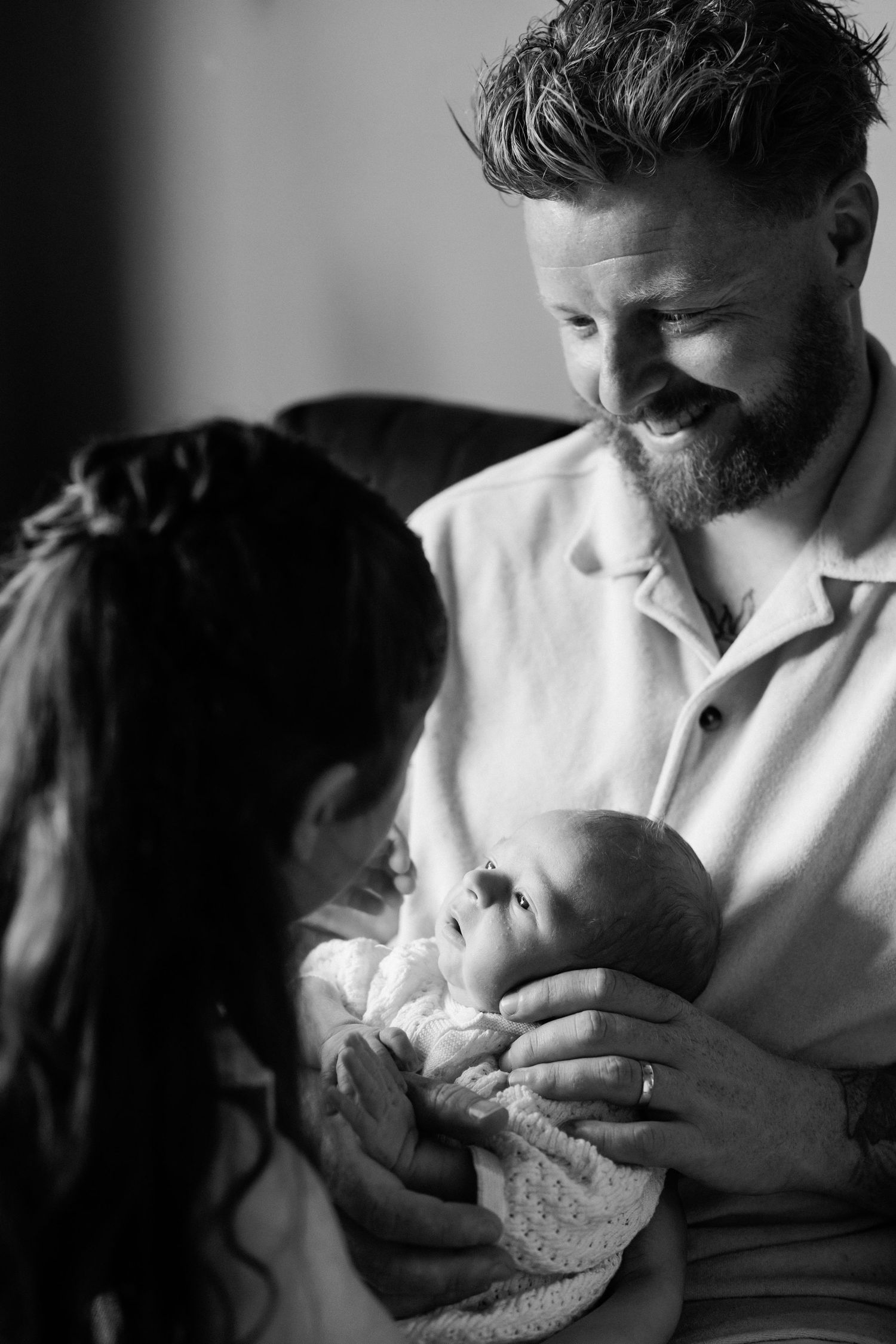 Black and white portrait of smiling bearded father and mother tenderly holding their newborn baby.