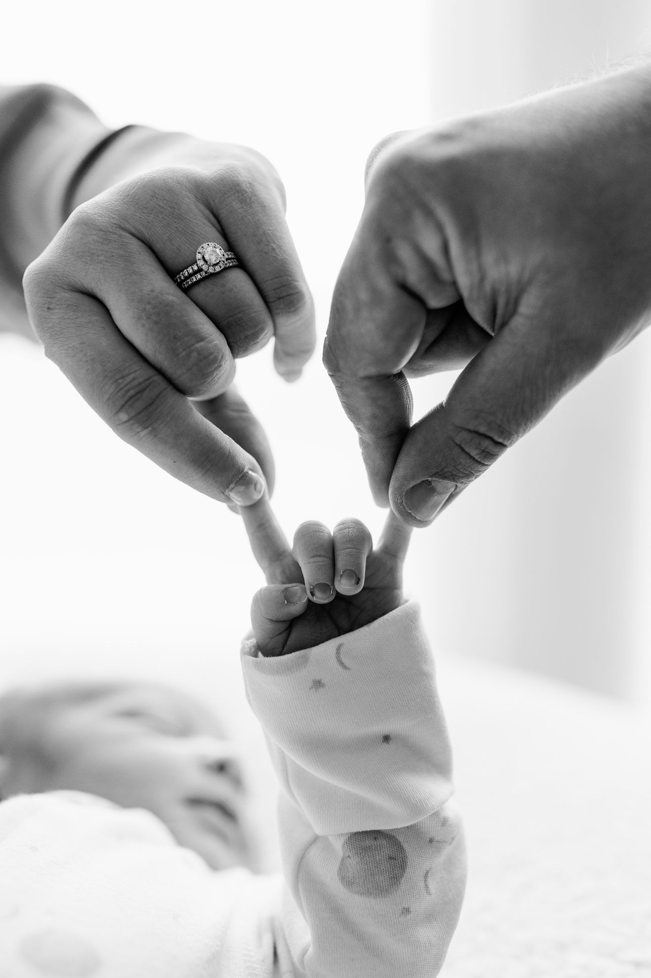 Black and white photo of two parents touching fingers with their sleeping newborn baby.