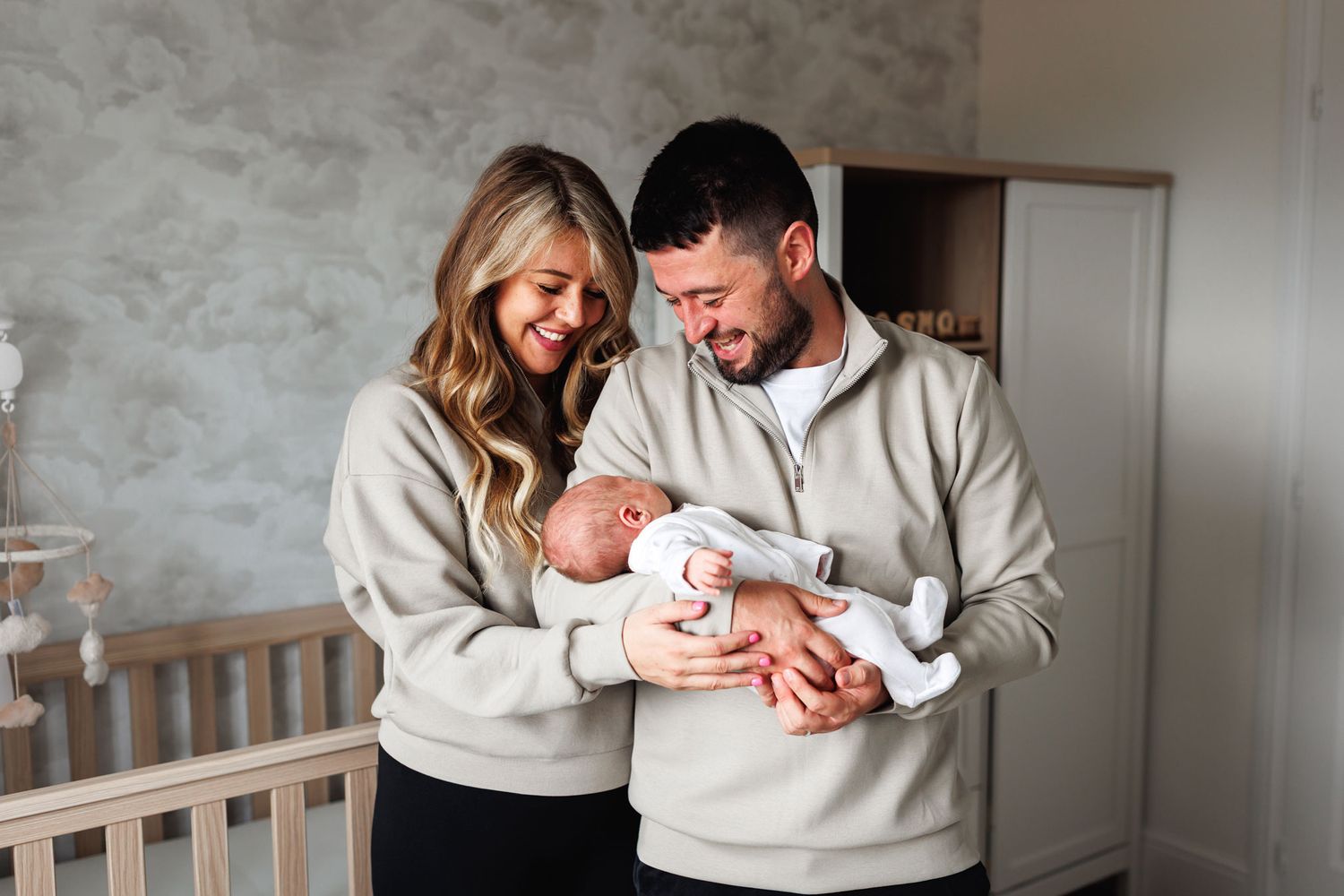 Happy new parents in matching grey sweatshirts smiling at their newborn baby in a decorated nursery.