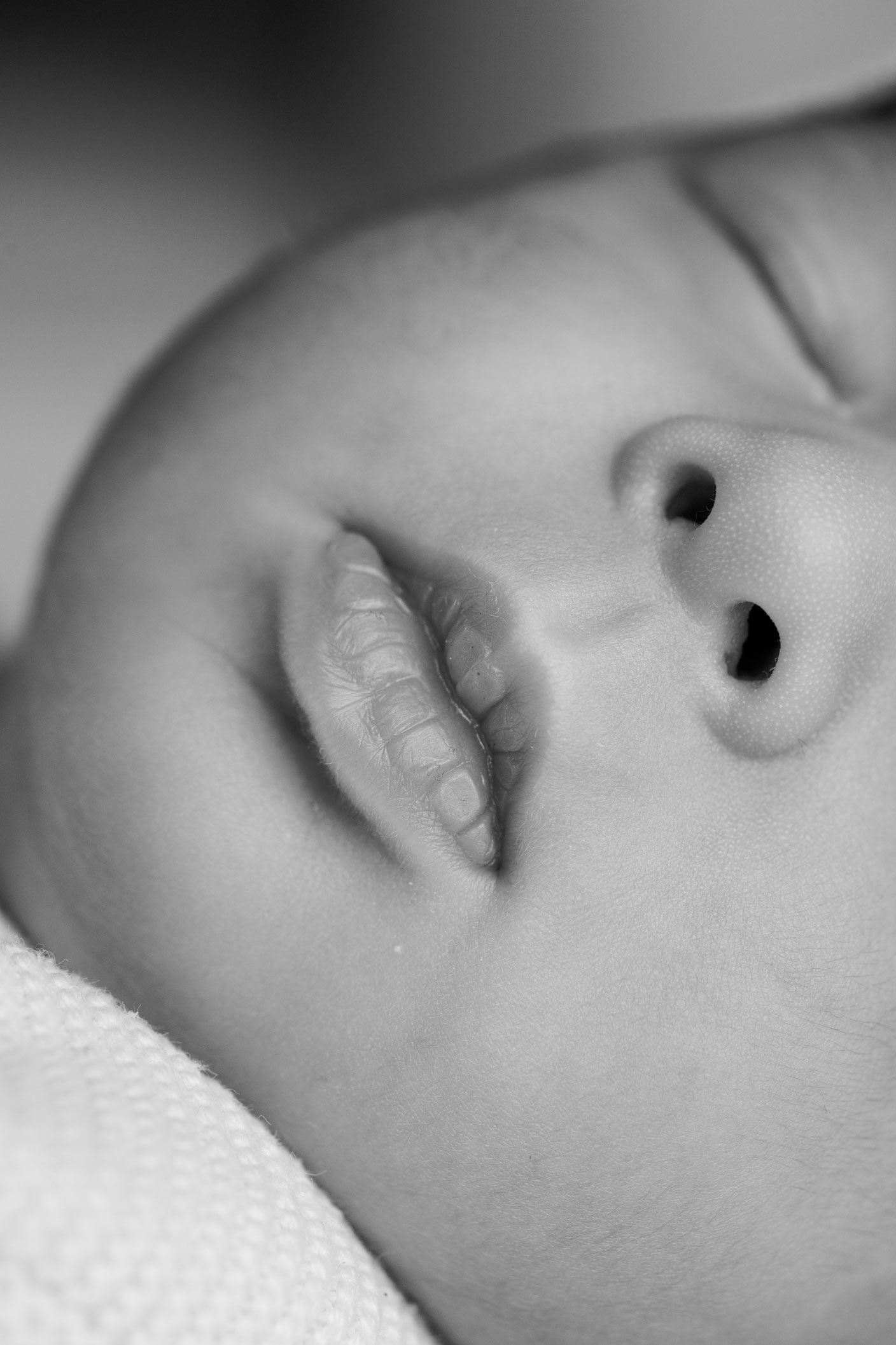Close-up black and white portrait of a sleeping newborn baby with soft cheeks, tiny nose and slightly parted lips resting on a knit blanket.