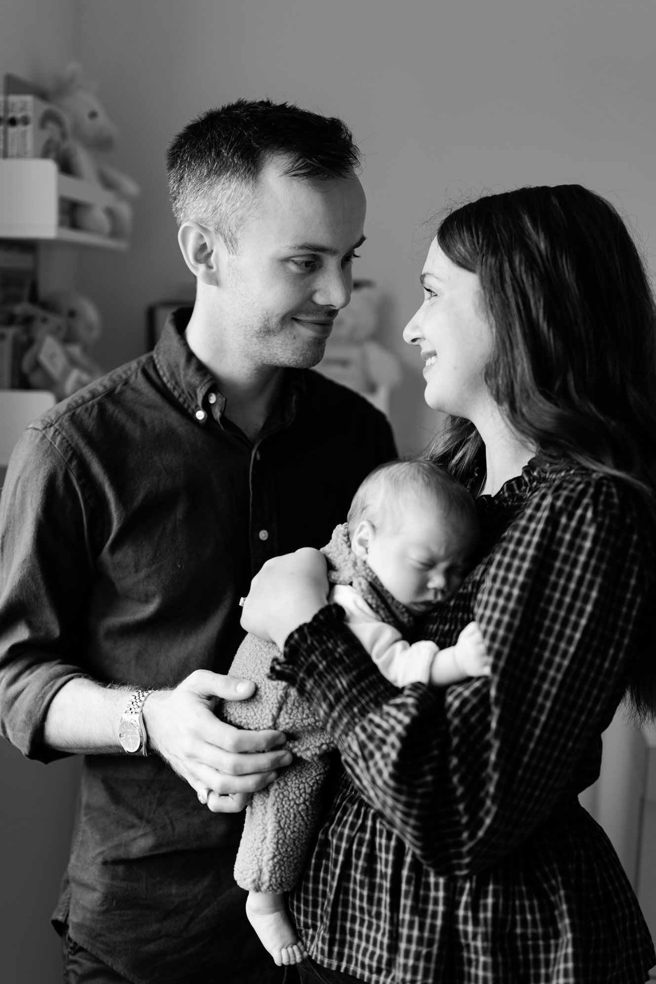 Black and white portrait of a smiling man and woman gazing at each other while holding a sleeping newborn baby in a cozy nursery.