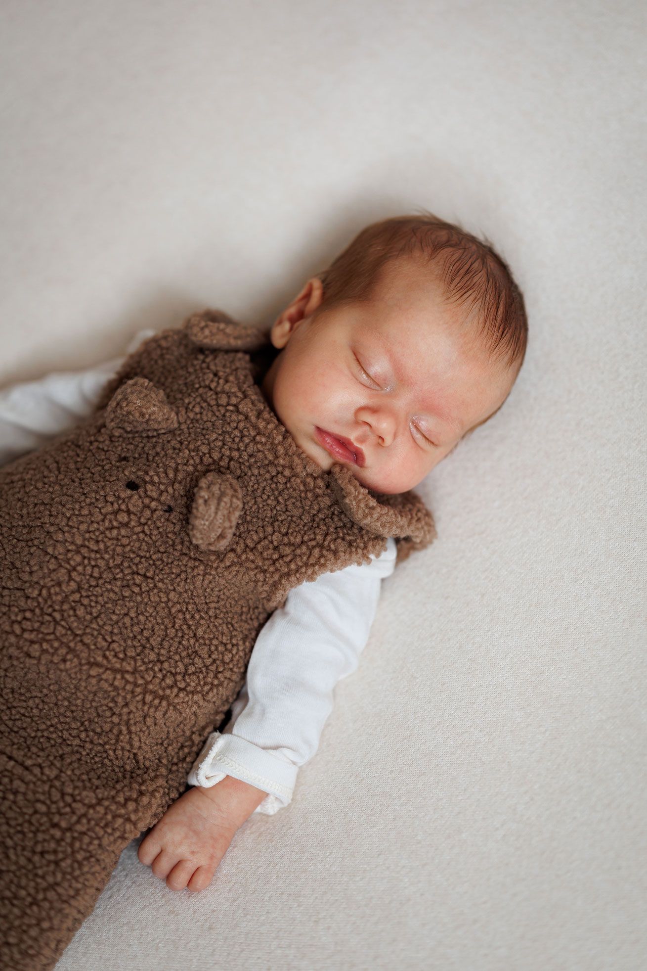 Sleeping newborn baby in a brown teddy bear fleece romper and white bodysuit lying peacefully on a soft cream blanket.