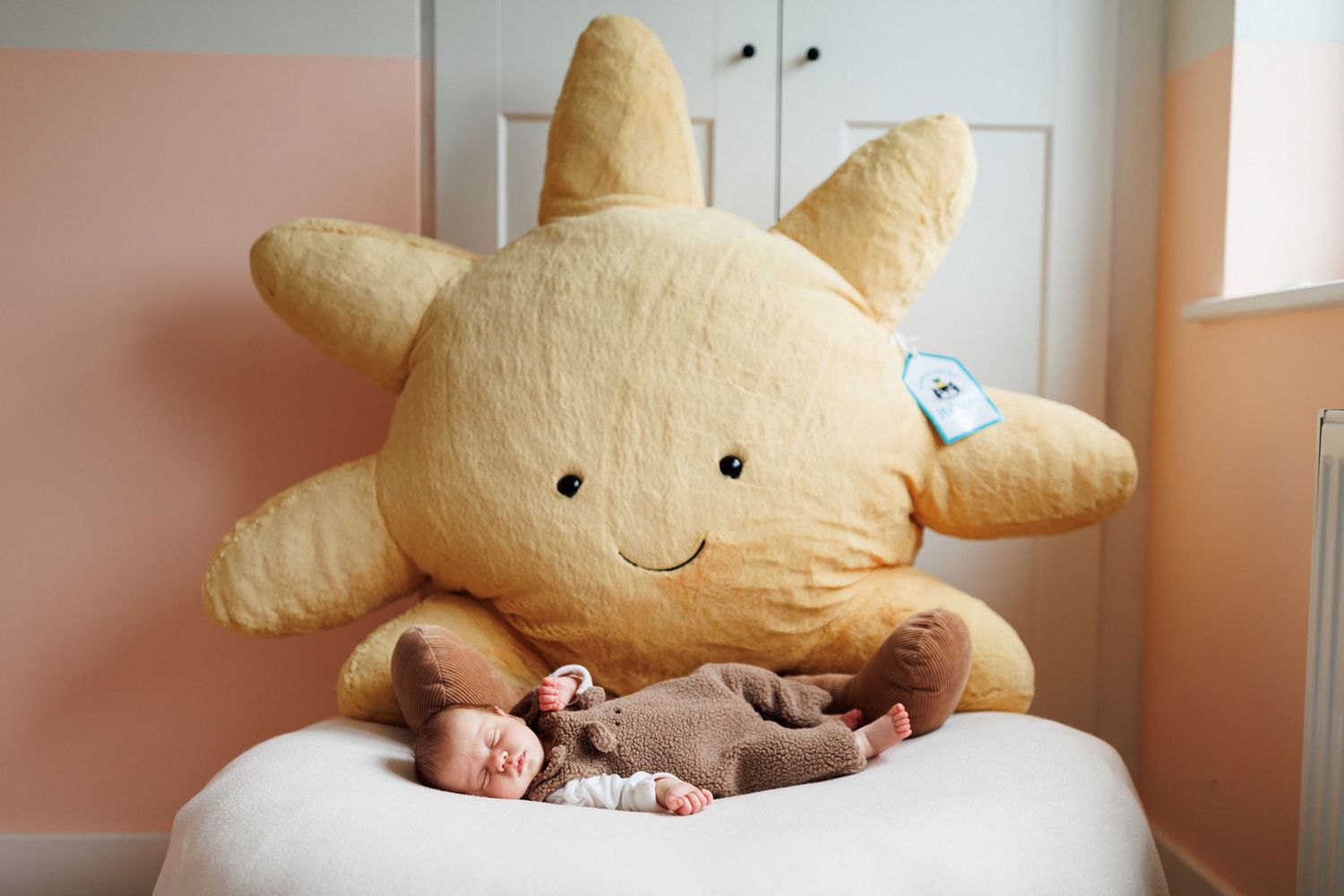 Newborn baby in a brown outfit sleeps on a white cushion beside a giant smiling sun plush pillow against a soft peach wall.