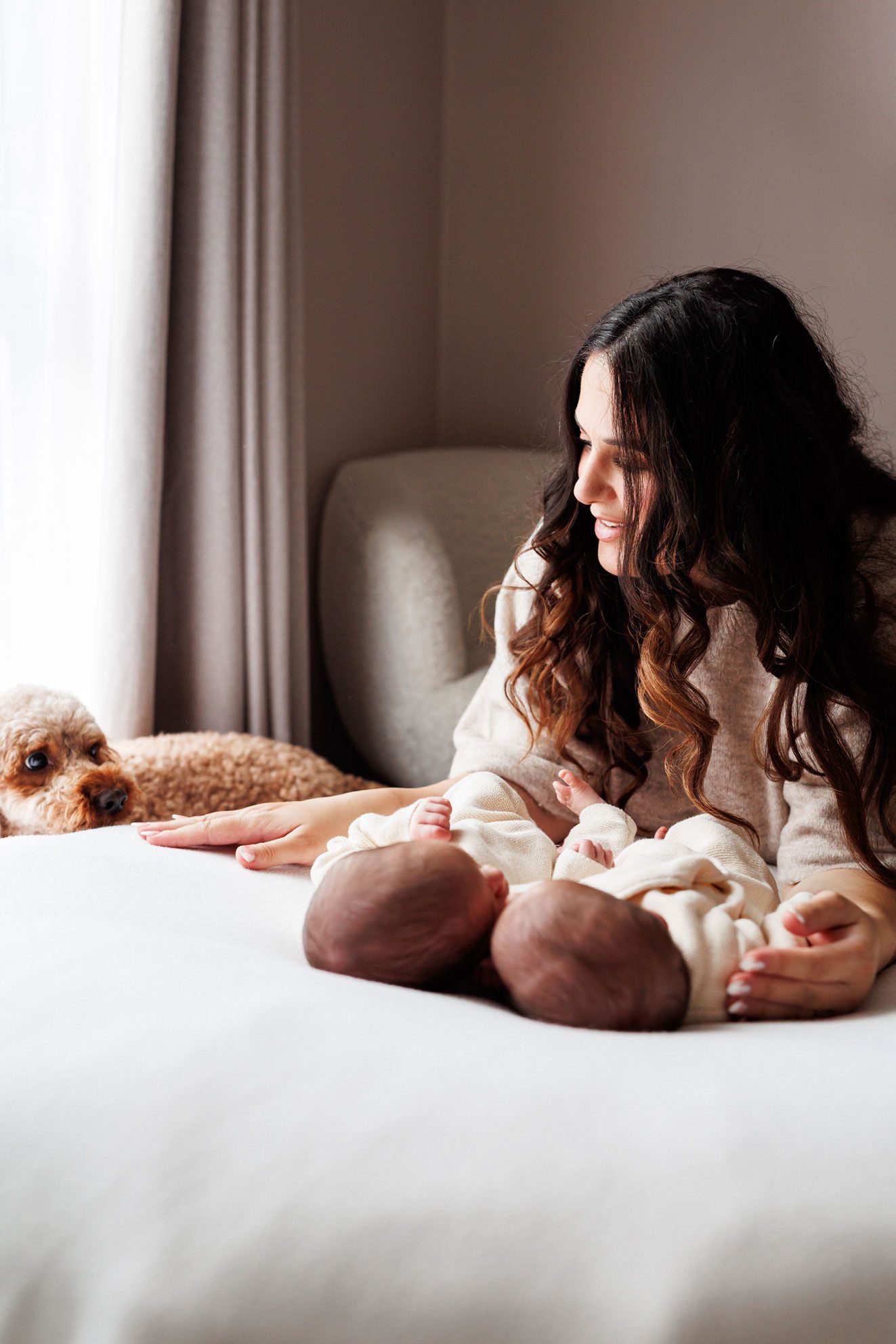 Smiling woman with long wavy hair sits on a white bed beside her newborn baby while a fluffy apricot dog rests nearby.