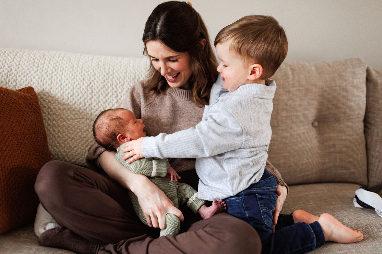 A smiling mother sits on a sofa holding her newborn while her toddler son gently touches the baby.