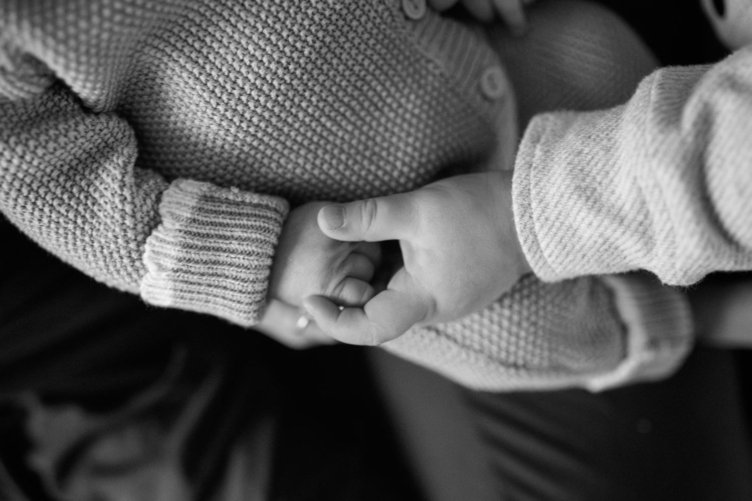 Black and white close-up of a newborn baby's tiny hand holding an adult's finger in a knit sleeve.