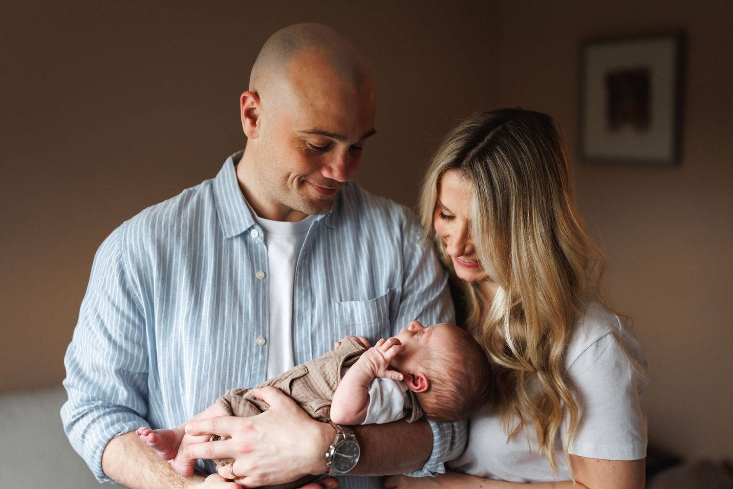 Happy parents smiling down at their newborn baby while holding them gently in a warm home setting in their Nottingham Newborn Photo Session