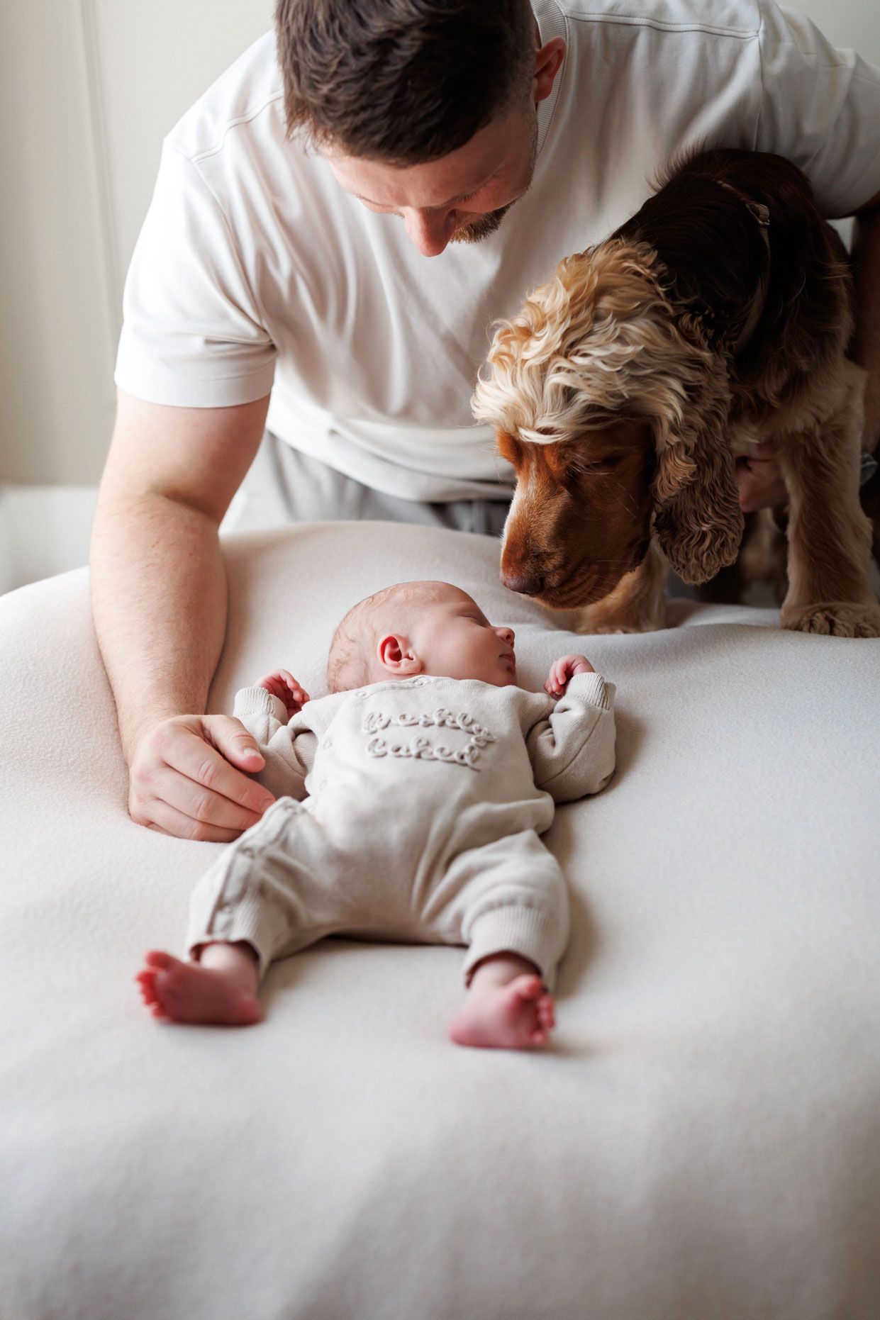 A man leans over a sleeping newborn baby lying on a cream blanket while a curious brown cocker spaniel sniffs the infant's head.