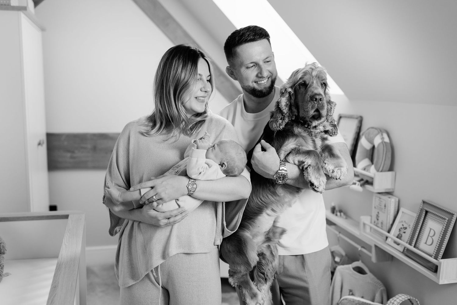 Smiling couple holding a newborn baby and a fluffy spaniel dog in a bright, cozy nursery in a black and white family portrait.