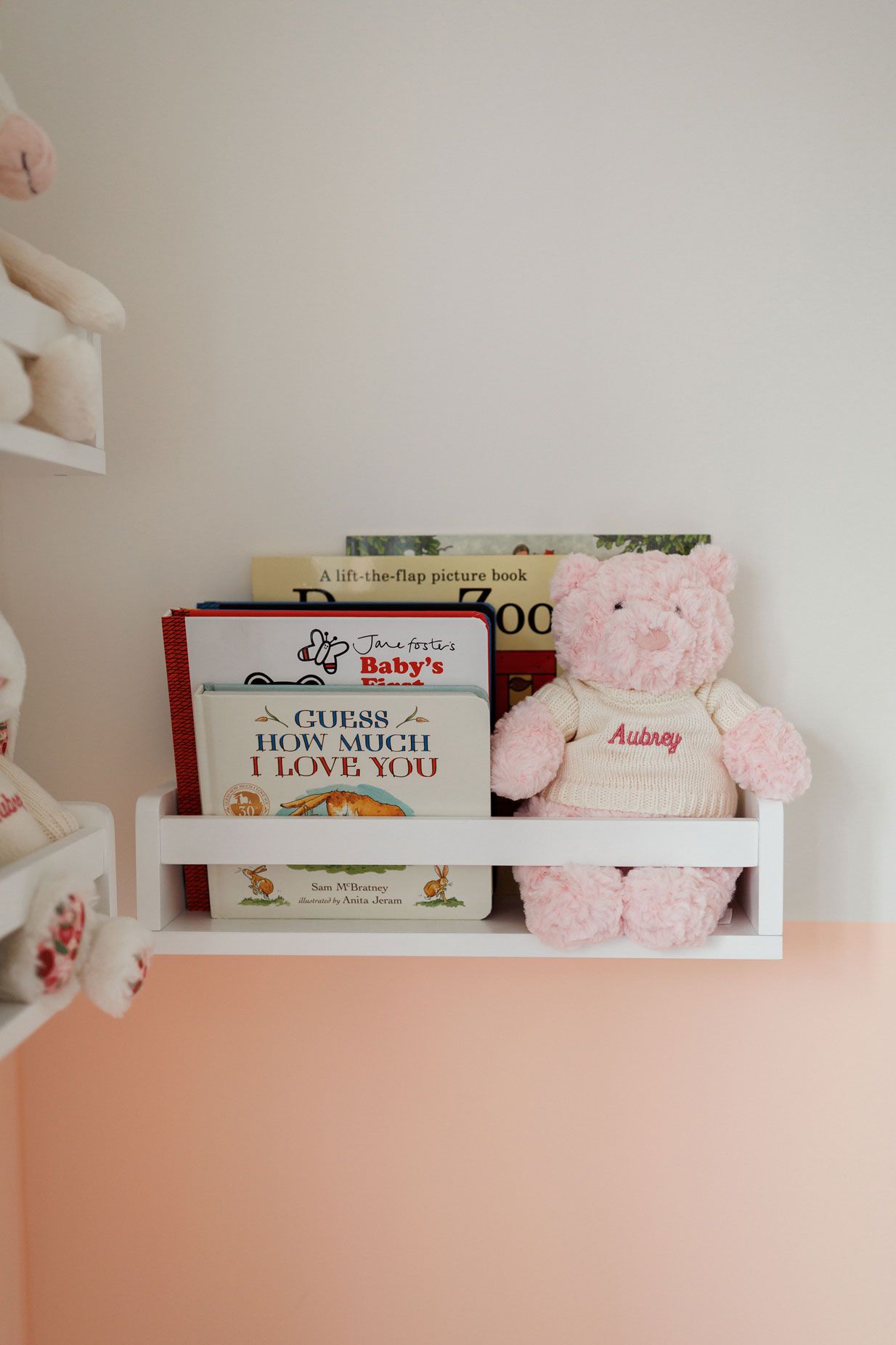 Pink teddy bear named Aubrey sits on a white nursery shelf beside children's books against a pastel pink wall.