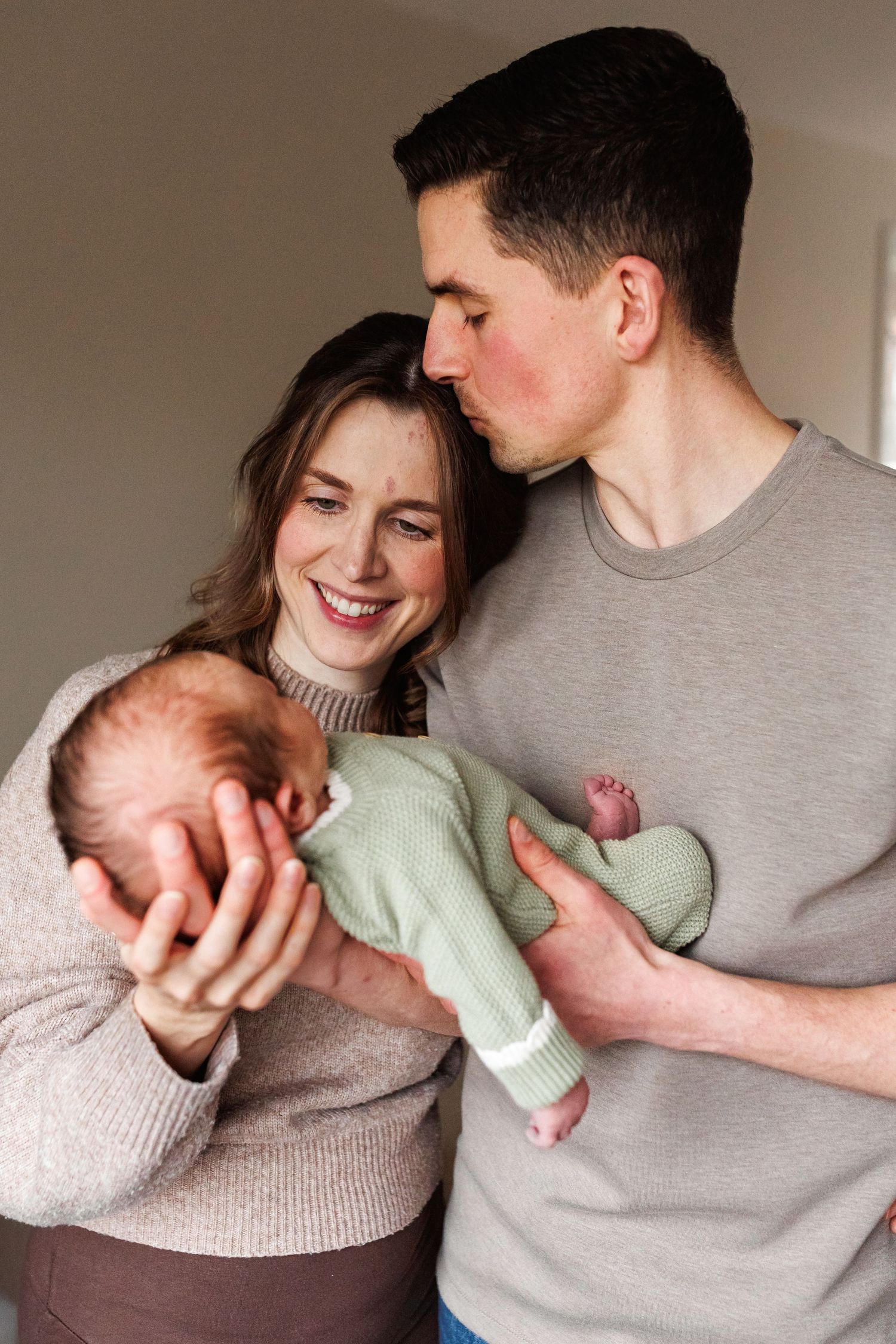 Happy new parents hold their newborn in a green knit outfit as the father kisses the mothers forehead.