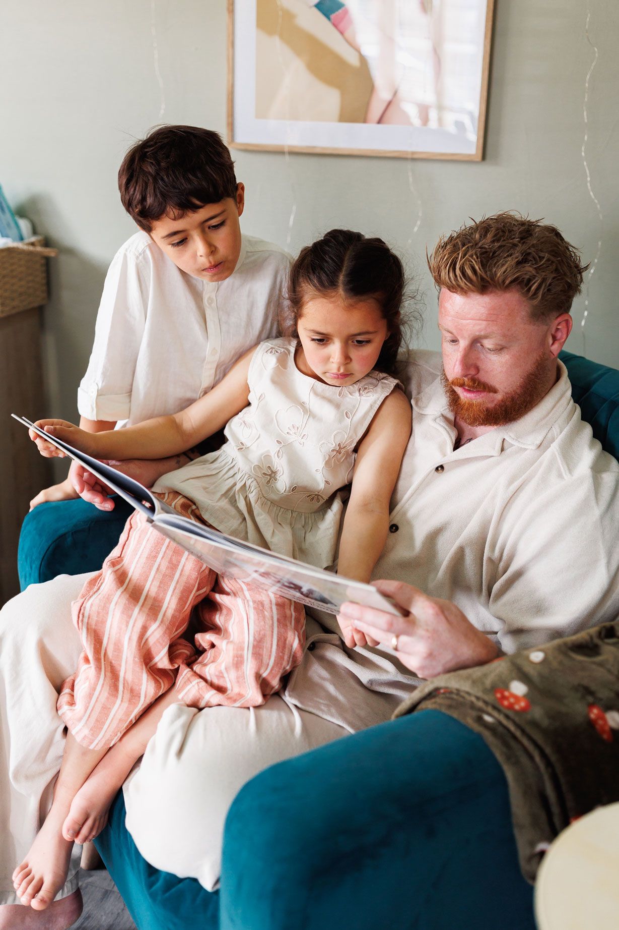 Red-bearded father reading a book with his two young children on a teal armchair at home.