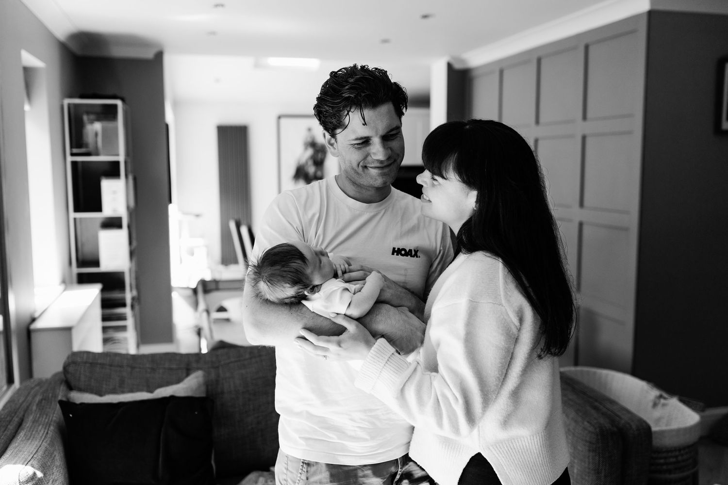 Black and white photo of parents smiling at their newborn baby in a cozy home setting.