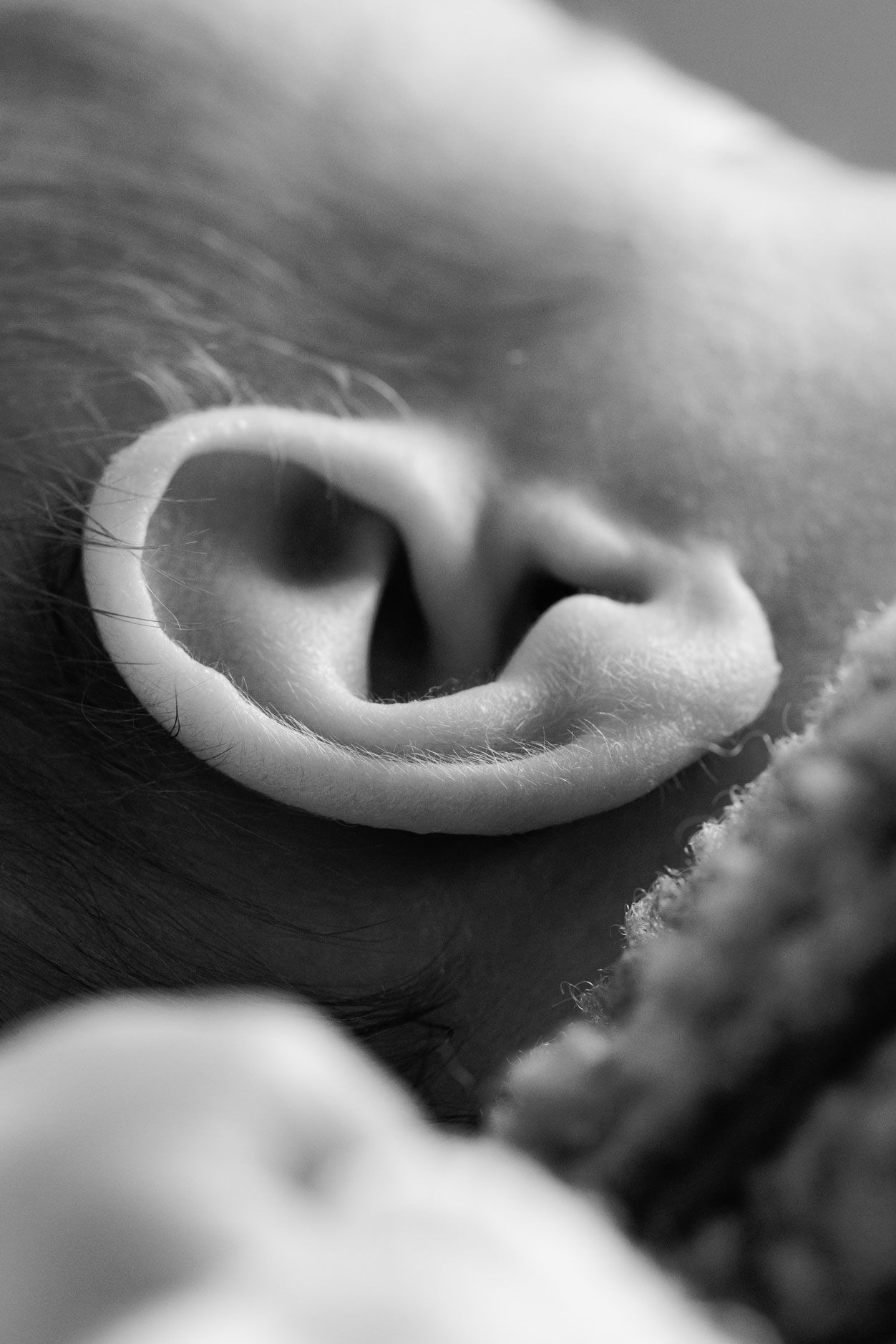 Black and white close-up of a sleeping baby's tiny ear, with soft downy hair and a textured knit blanket blurred in the foreground.