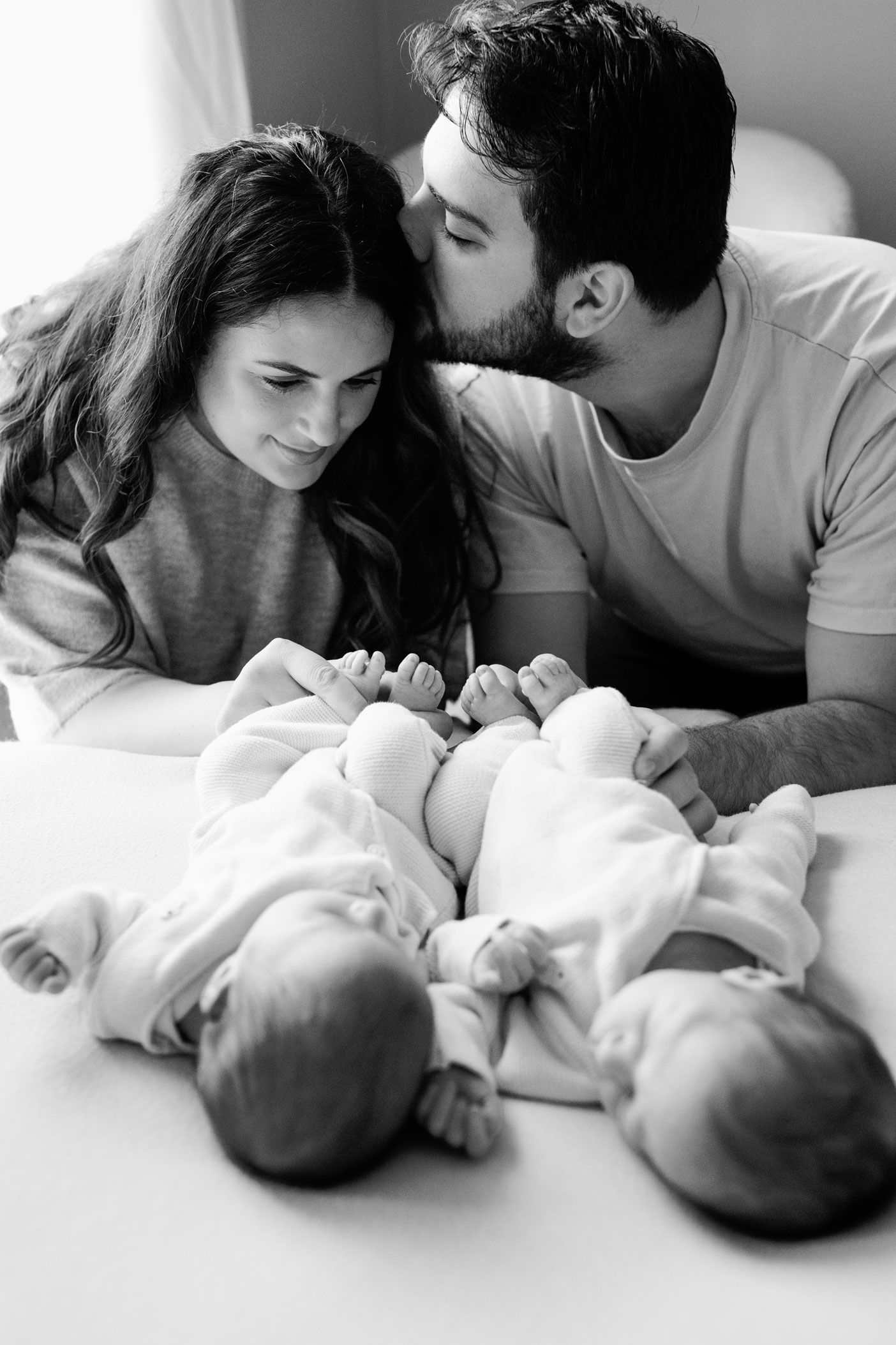 Black and white photo of a smiling mother and father leaning over two newborn twin babies lying side by side on a bed.