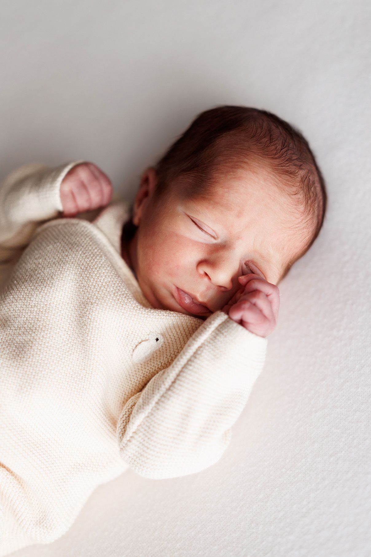 Sleeping newborn baby in a cream knit outfit lying on a soft white blanket with tiny hands raised near the face.