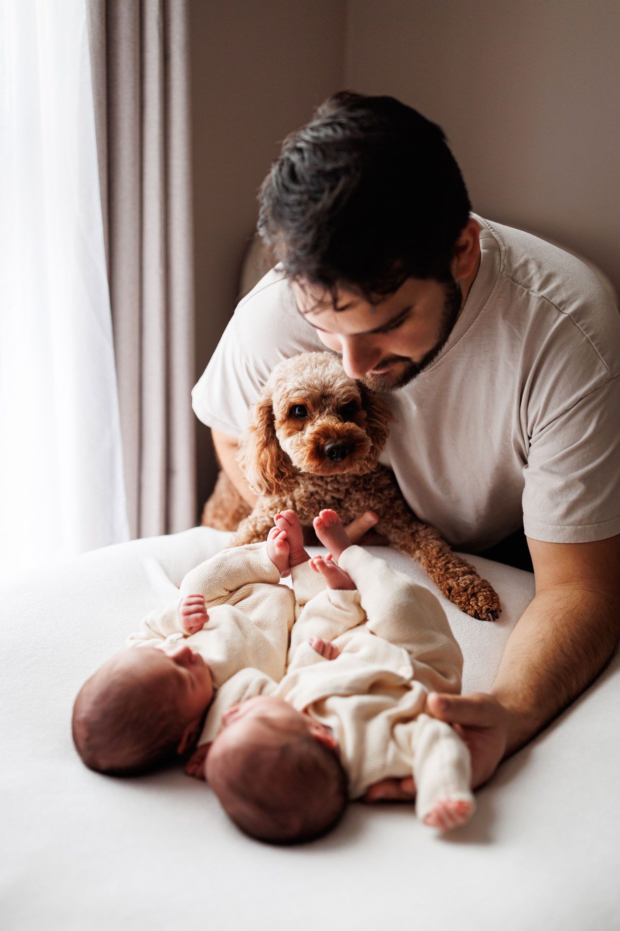 A man in a white t-shirt holds a fluffy apricot poodle above two sleeping newborn babies in cream outfits on a bed by a sunlit window.