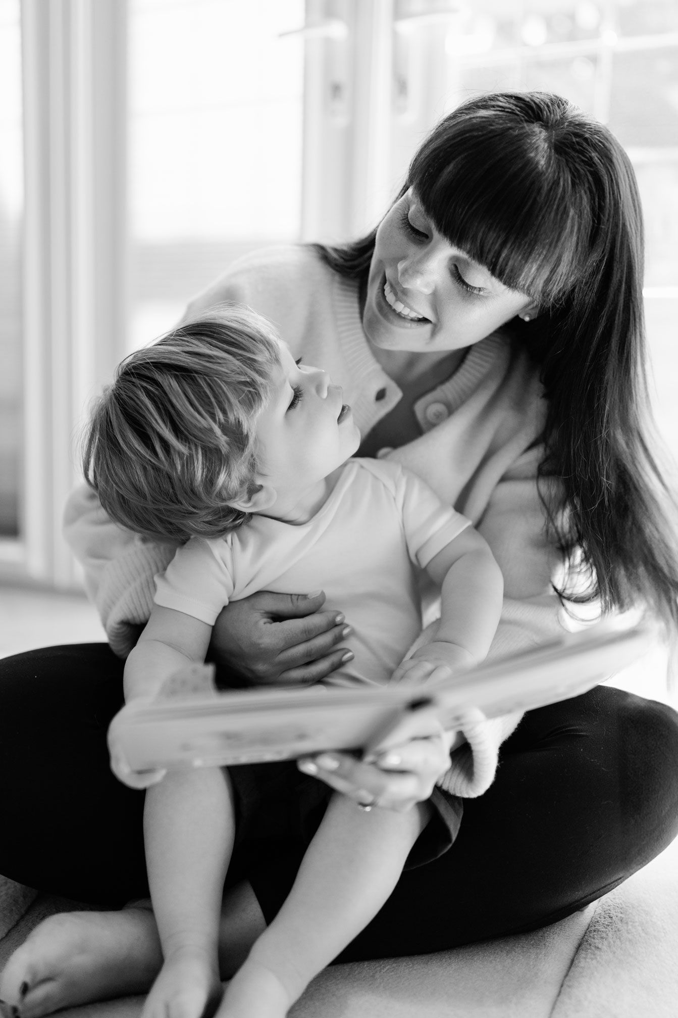Black and white photo of a smiling mother holding her toddler while reading a book together at home.