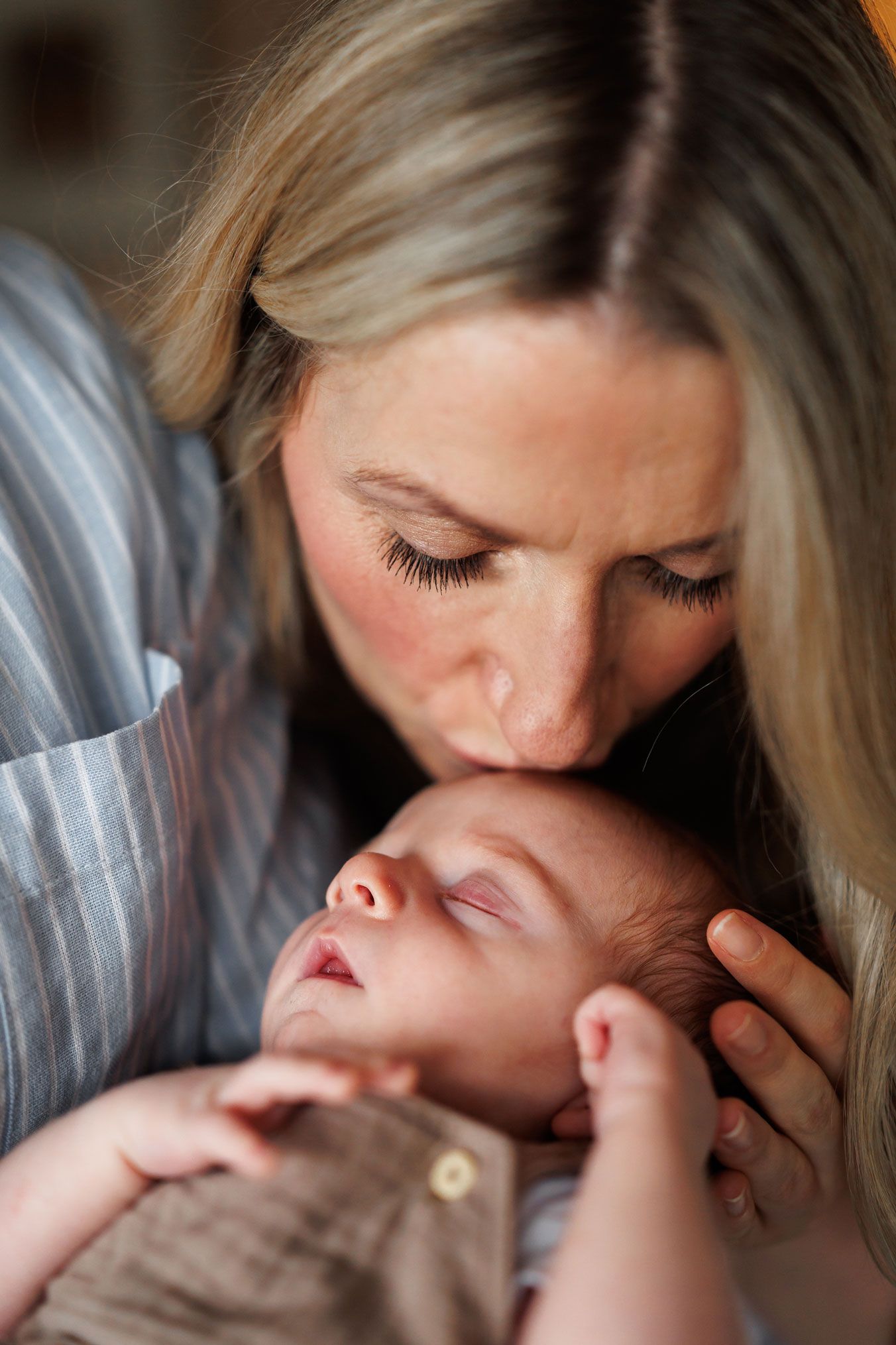 A tender moment as a blonde mother gently kisses her sleeping newborn baby on the forehead.