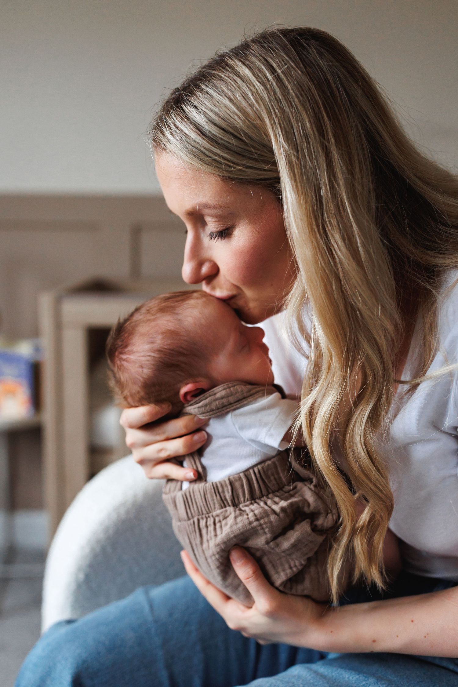 Blonde mother in white shirt gently kissing her newborn baby on the head while seated.